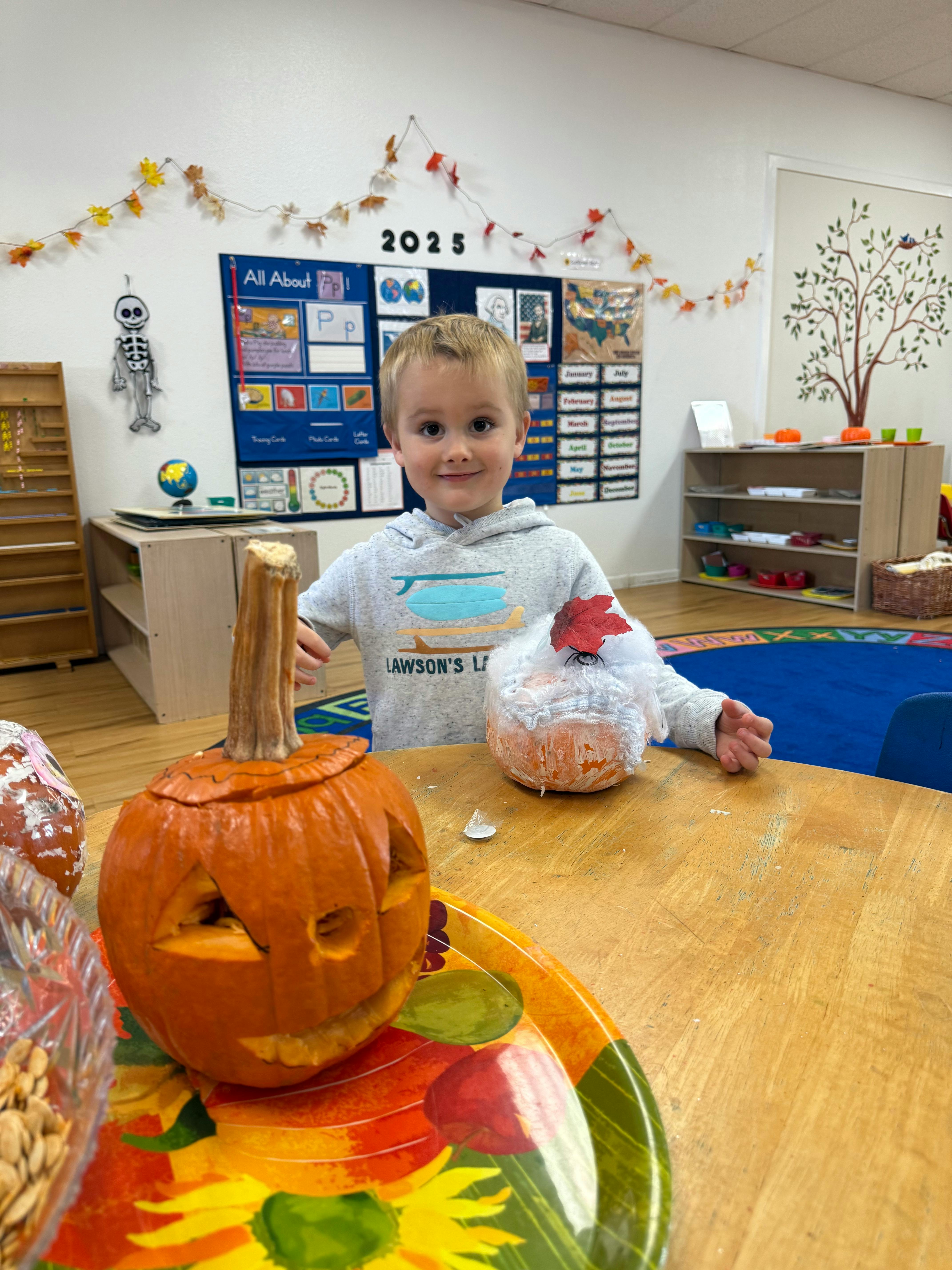Child standing behind a carved pumpkin and a pumpkin craft in a classroom