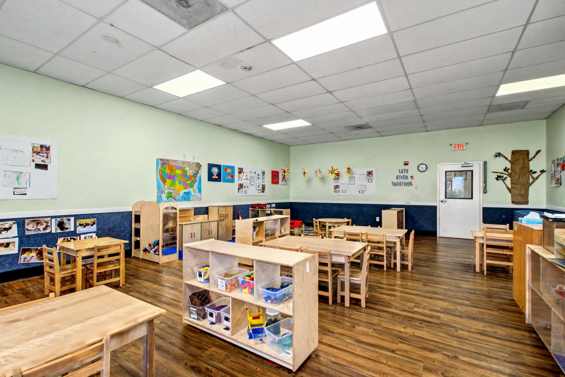 Empty classroom with small wooden tables and chairs, shelves with toys