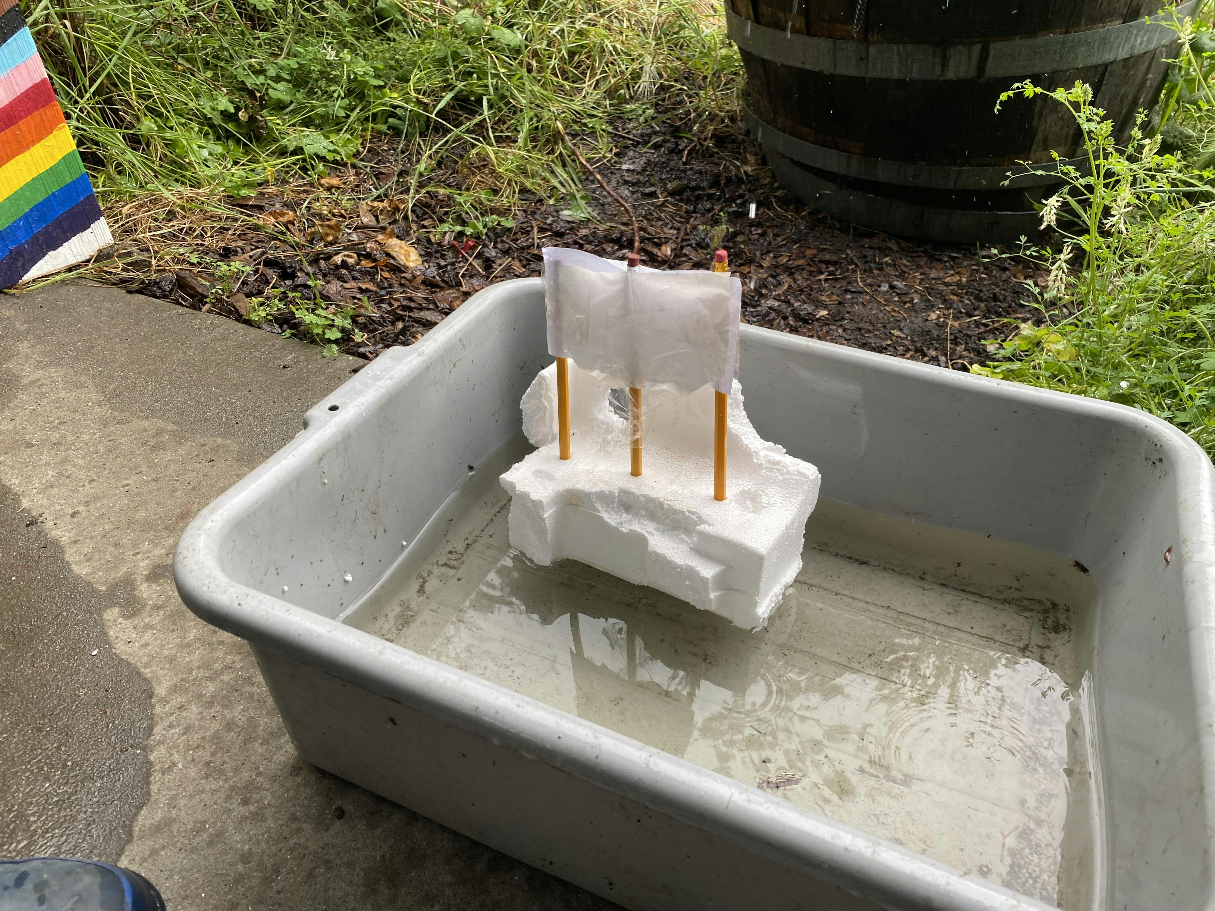 A white styrofoam boat with pencil masts and paper sails floating in a tub of water