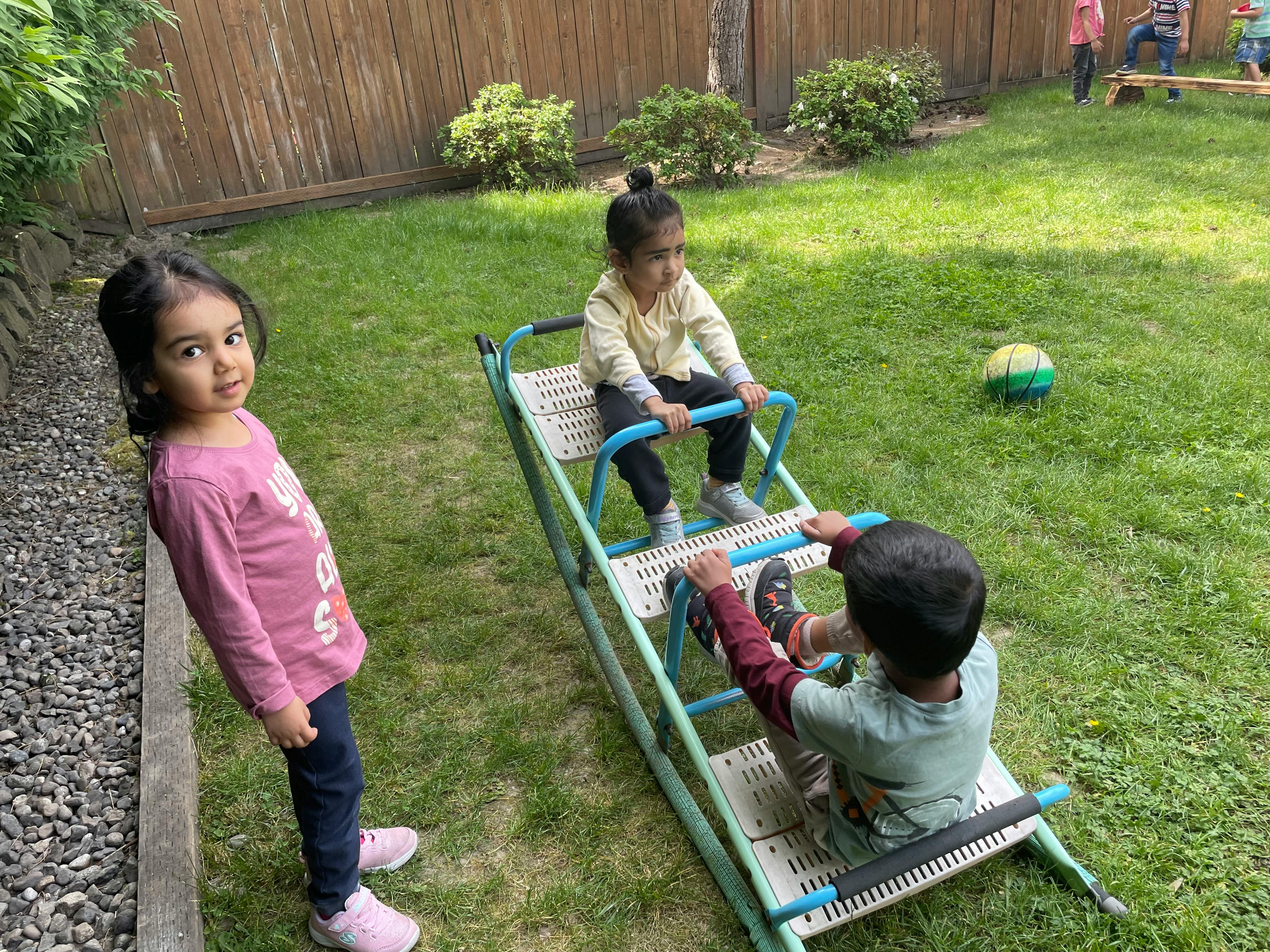 Two children on a seesaw and one child standing nearby in a grassy area