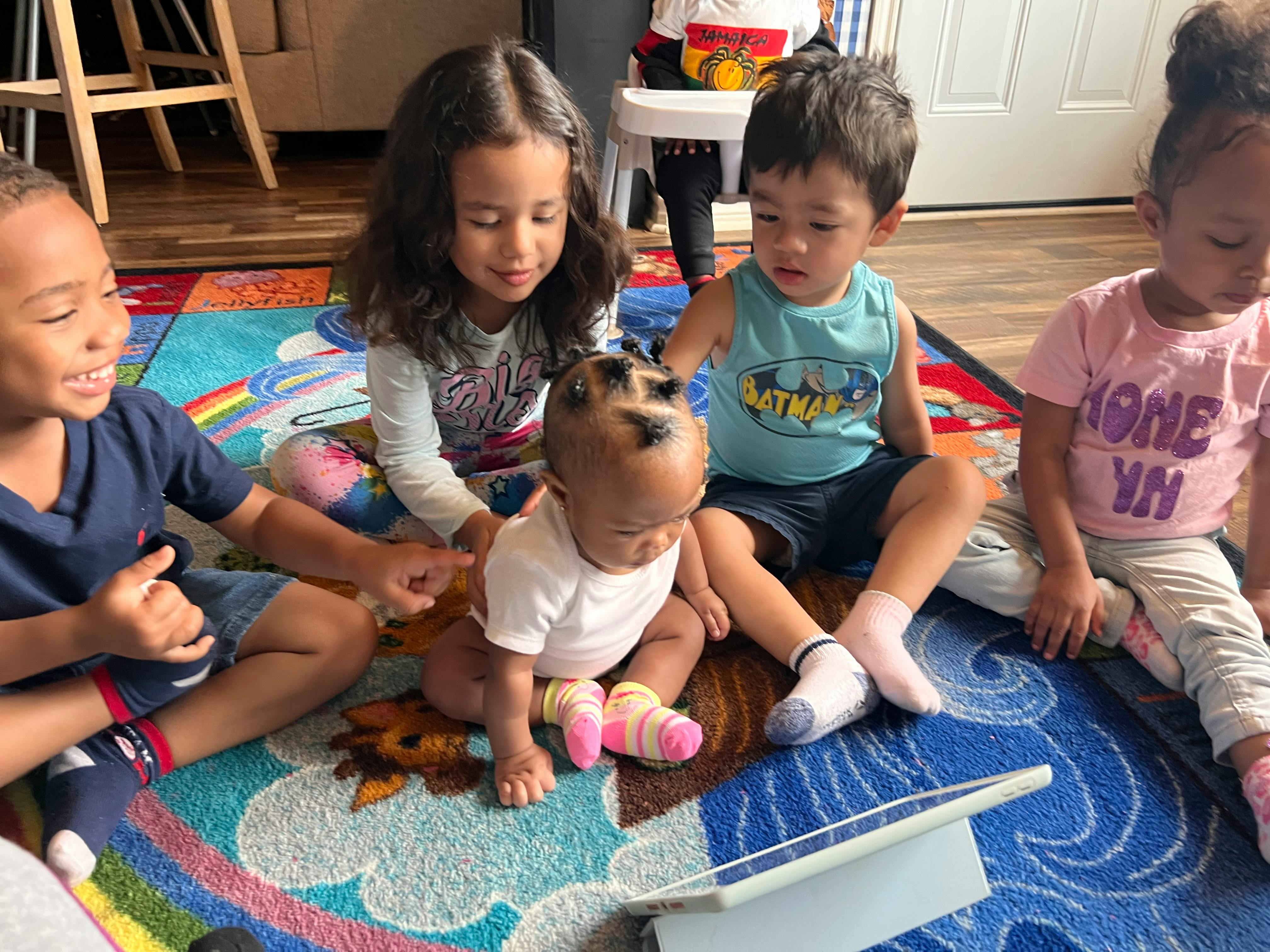 Four children and one baby sitting on a colorful rug looking at a tablet
