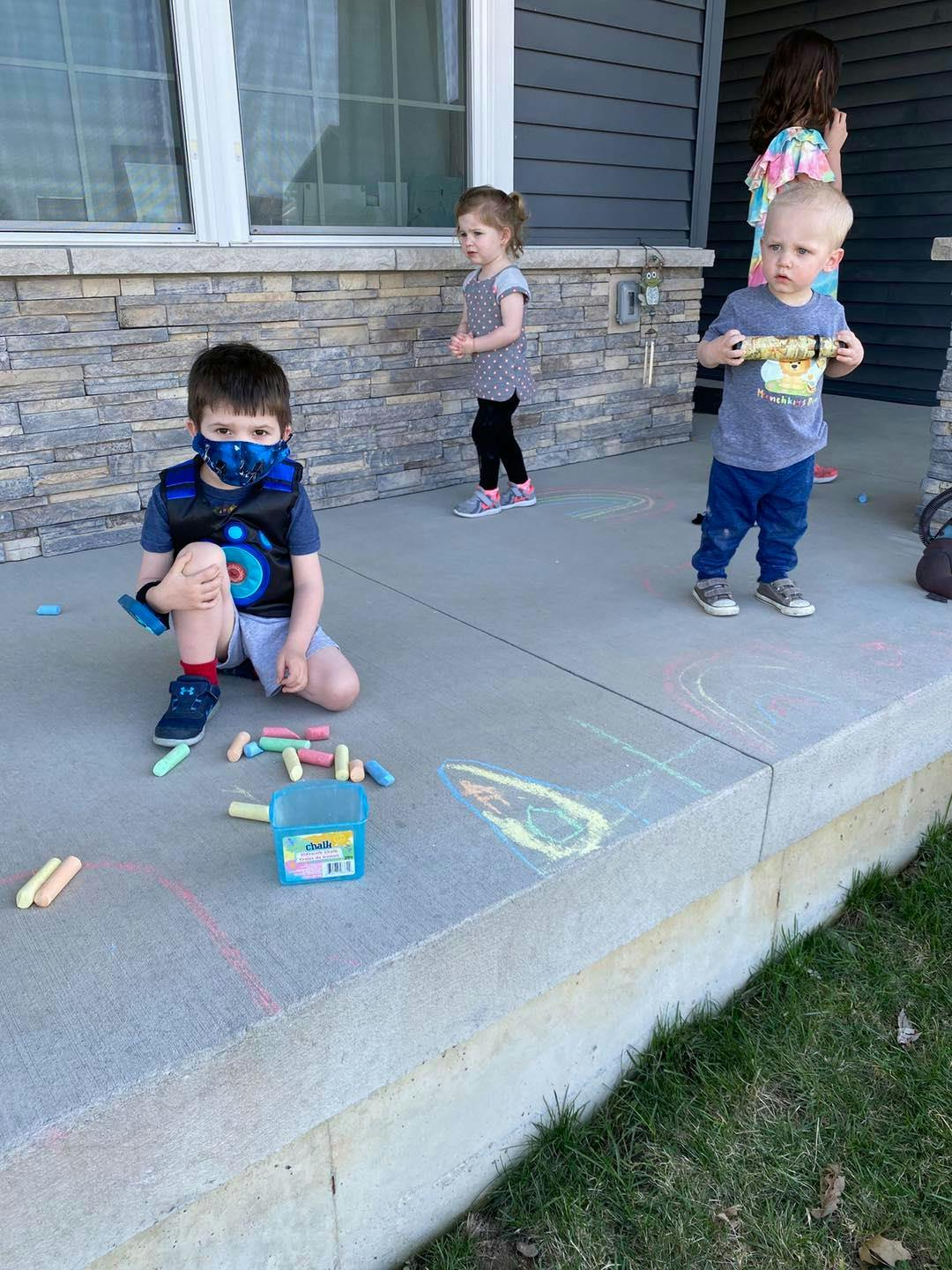 Child in mask kneeling on porch with chalk, two other children standing