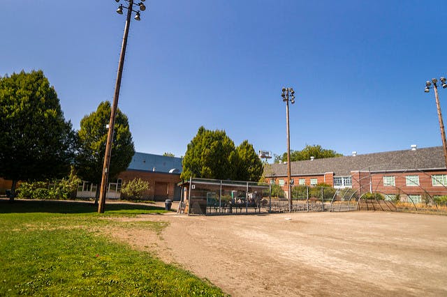 Dirt baseball field with bleachers and brick buildings in the background