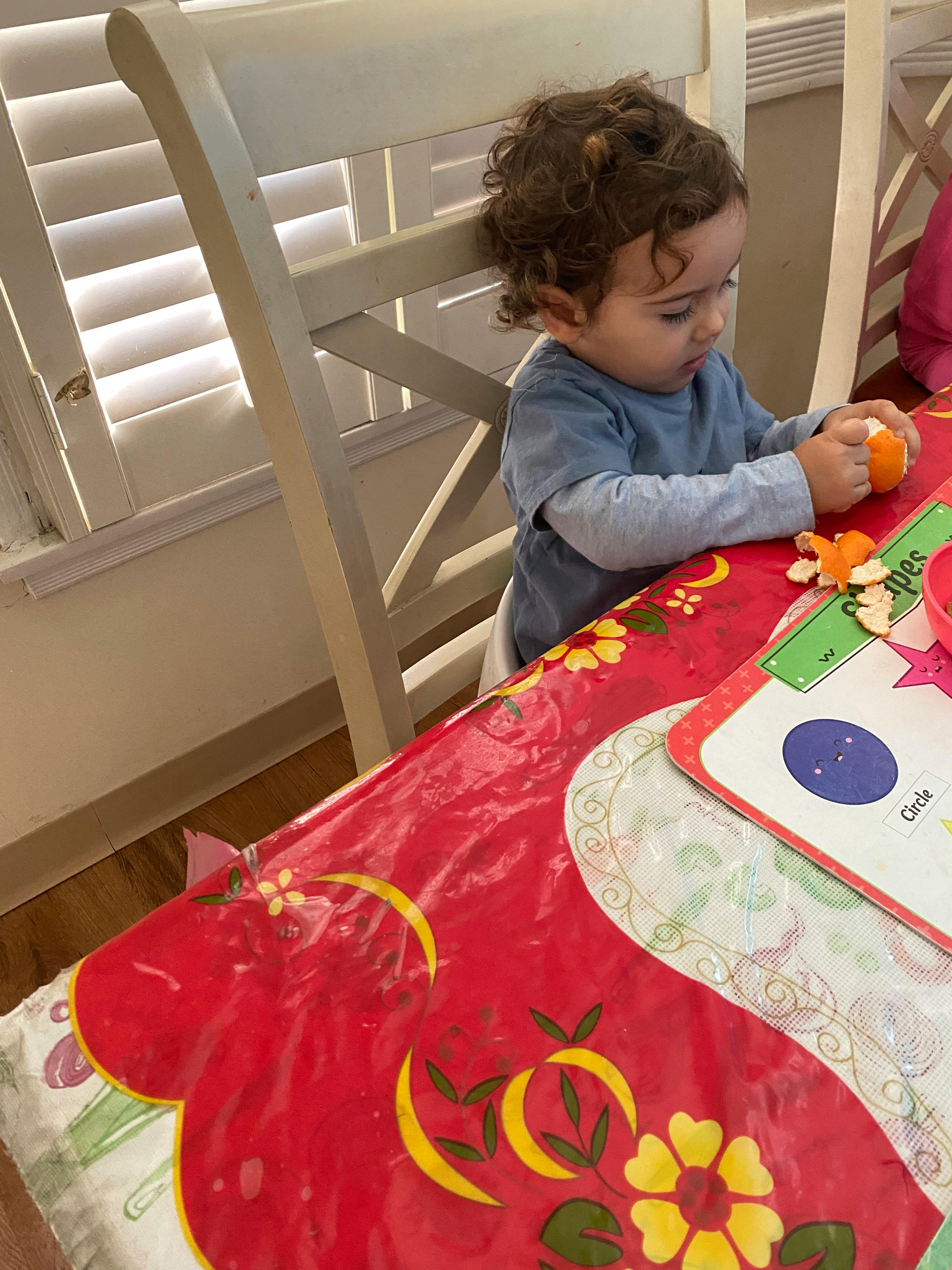 Child sitting at a table with a red tablecloth, holding an orange object