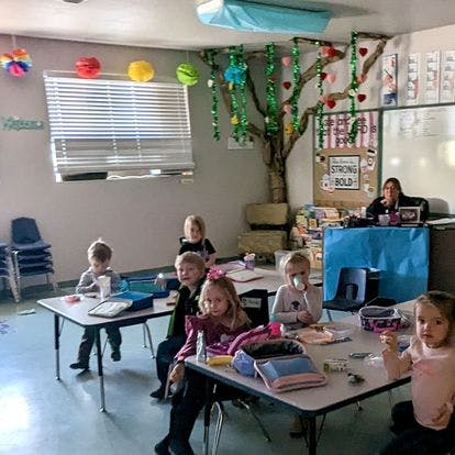 Children sitting at tables in a classroom with an adult at a desk