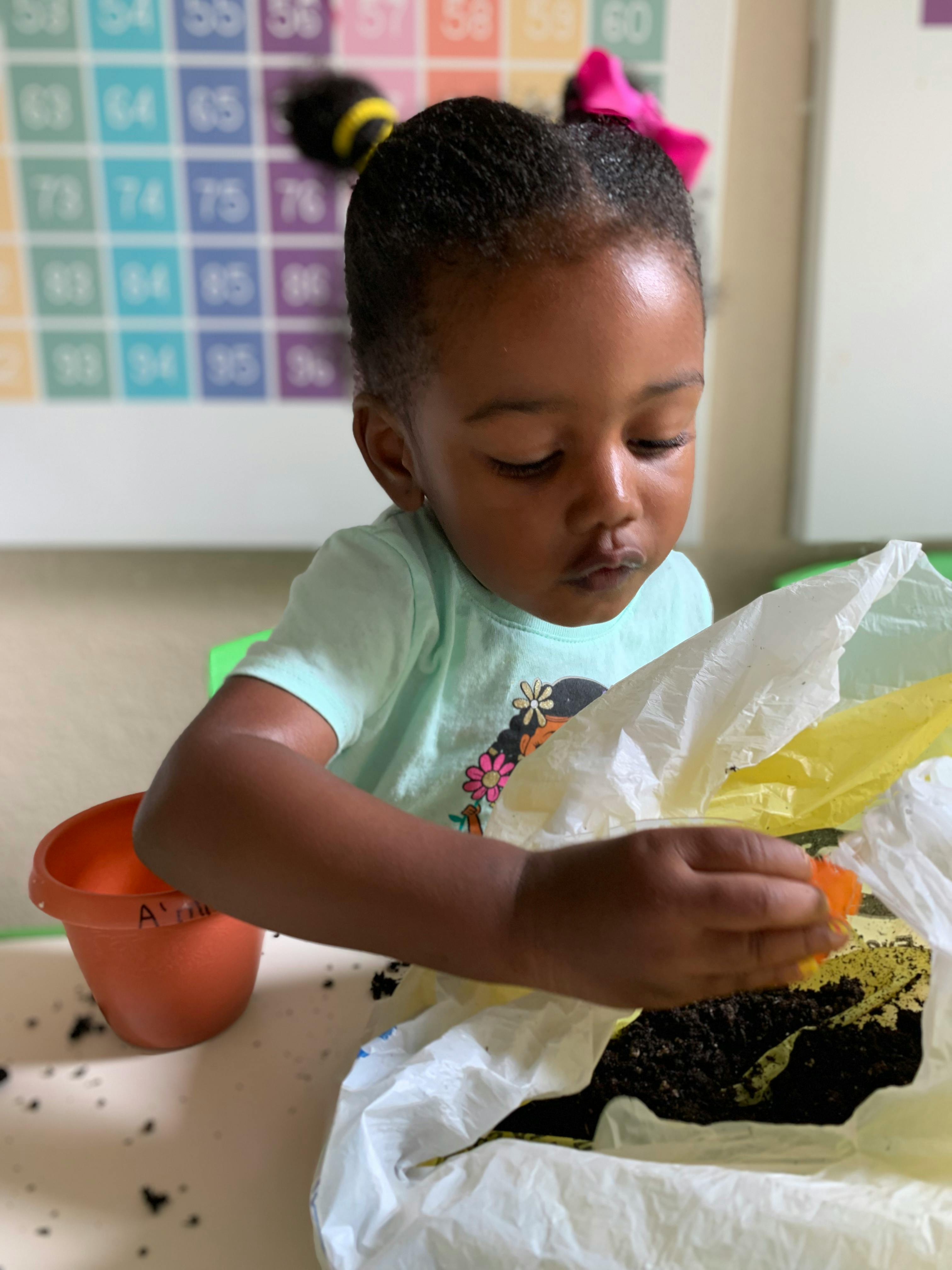 Child holding a small orange object over a bag of soil