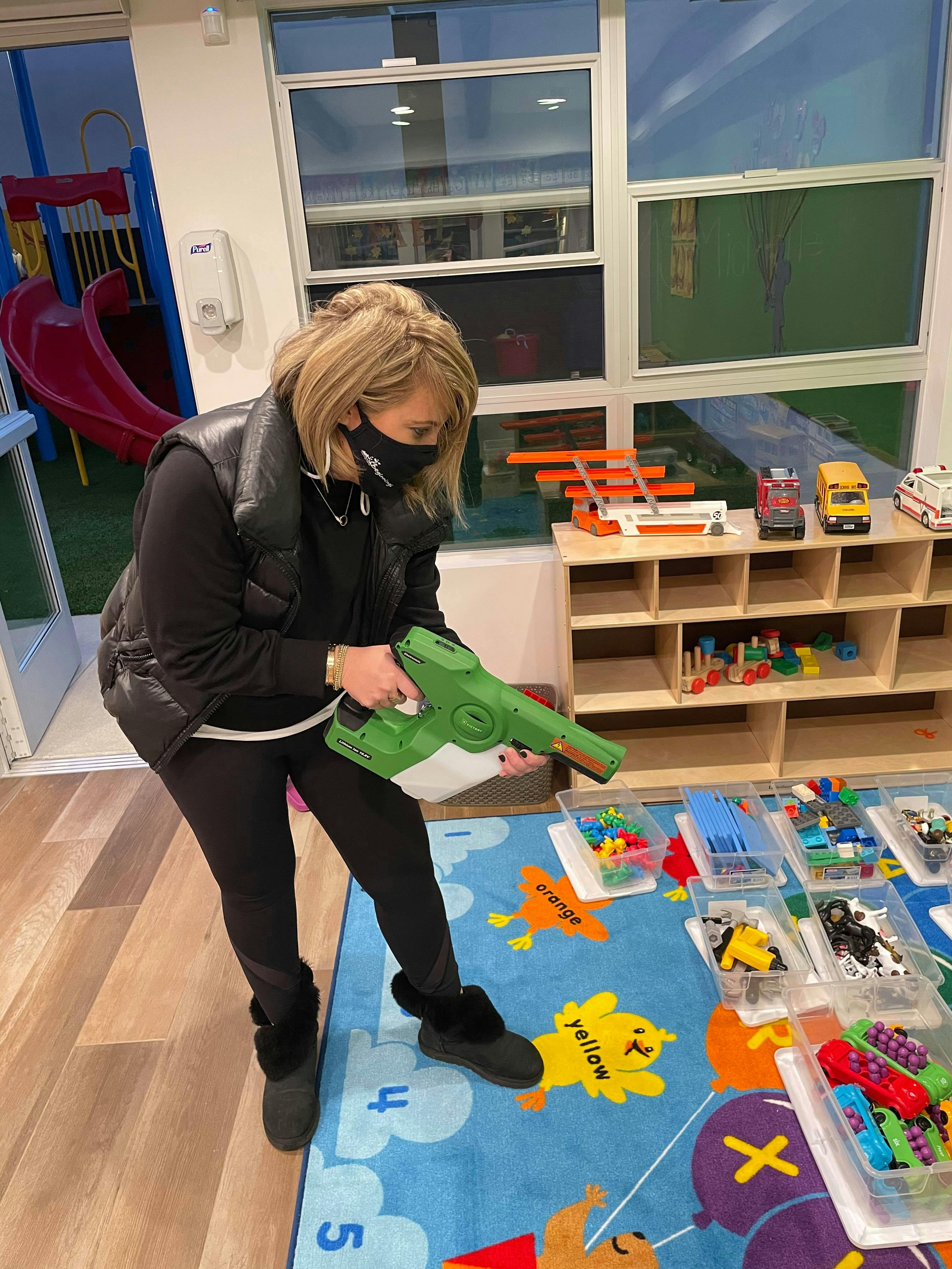 Person in a mask holding a green and white sprayer in a room with toys