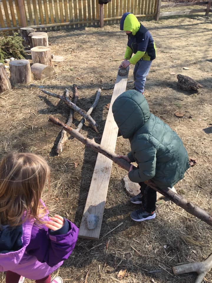 Three children playing with sticks and a wooden plank outdoors