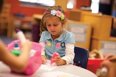 Child at a table holding a pink object and looking down at a paper