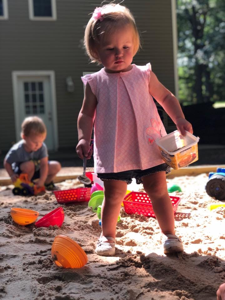 Child standing in sandbox holding a container, another child playing in background