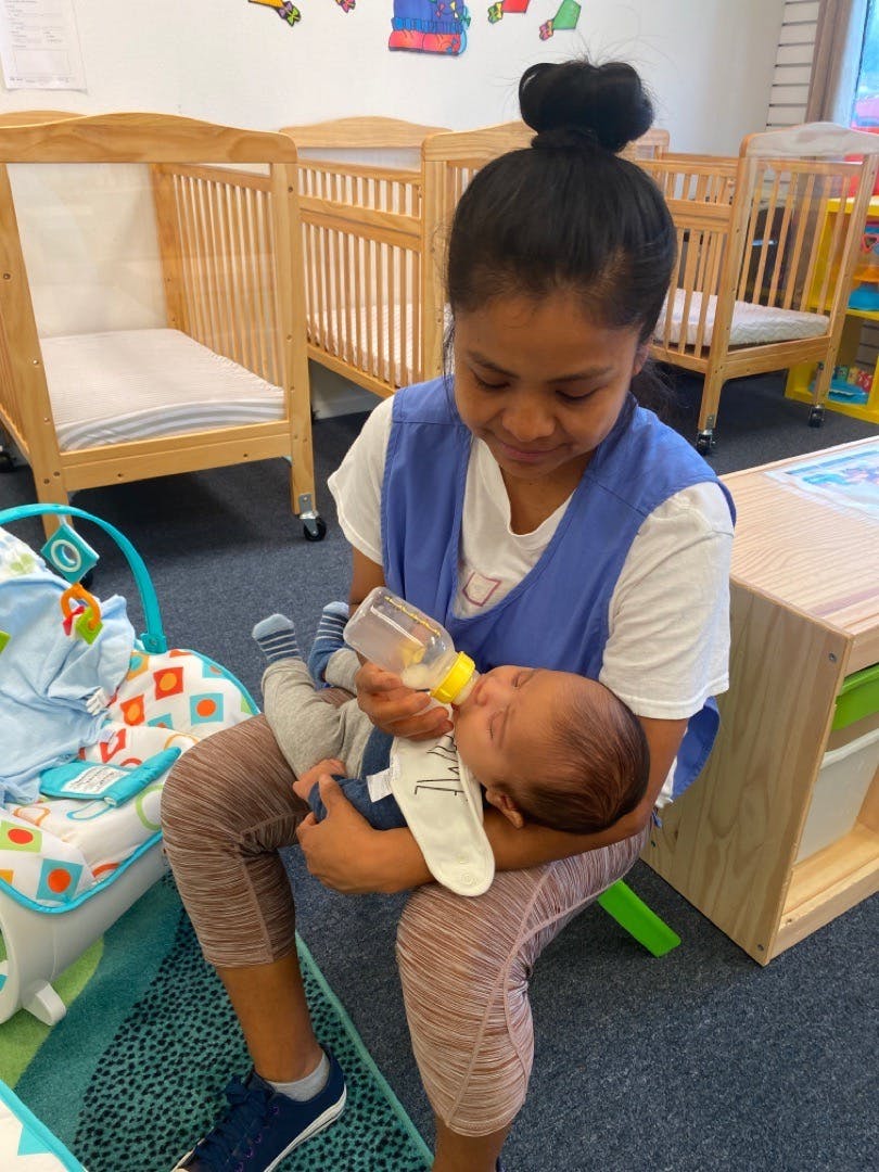 Person feeding a baby with a bottle in a room with cribs