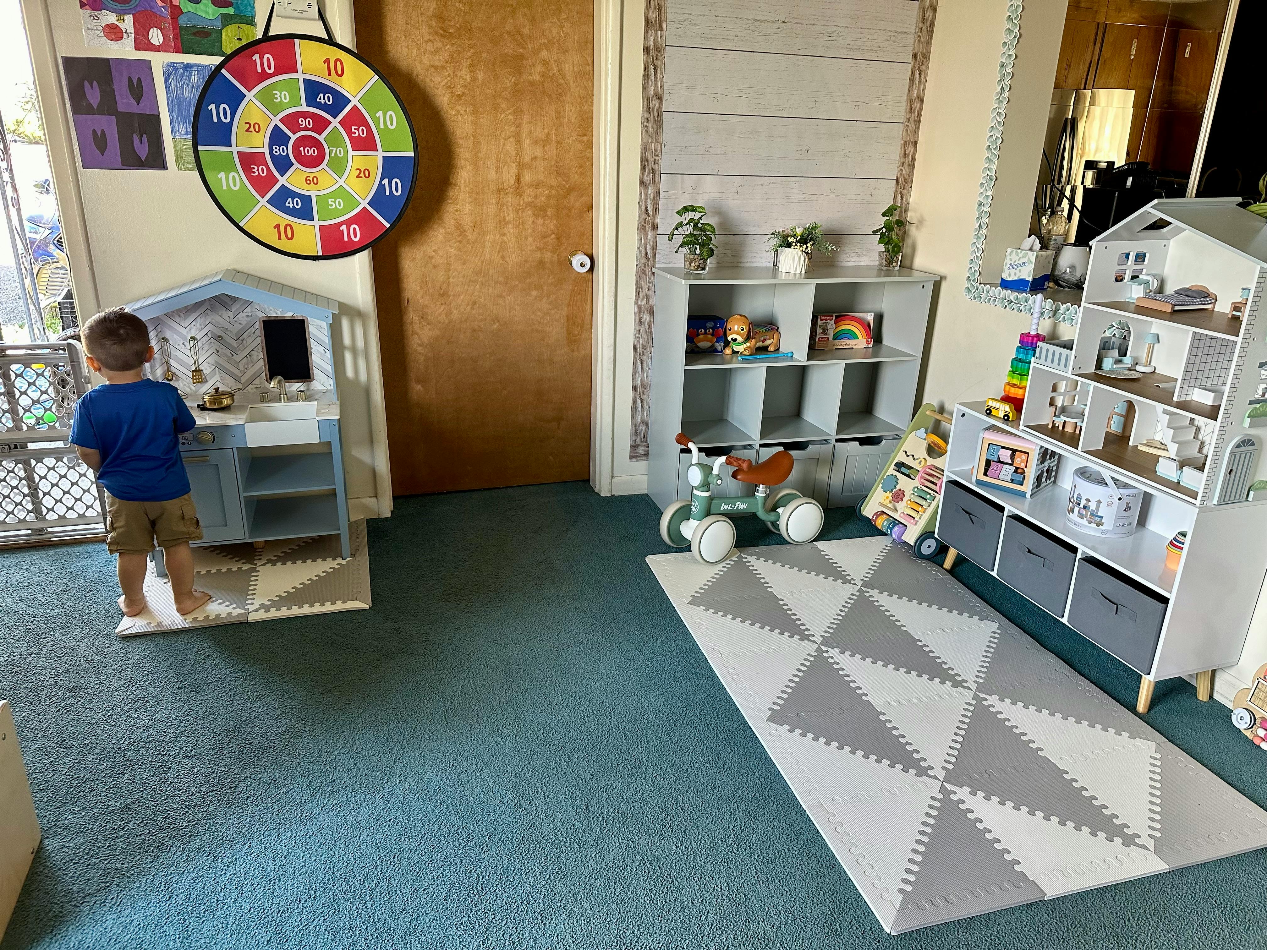 Child standing next to a play kitchen in a room with toys and shelves