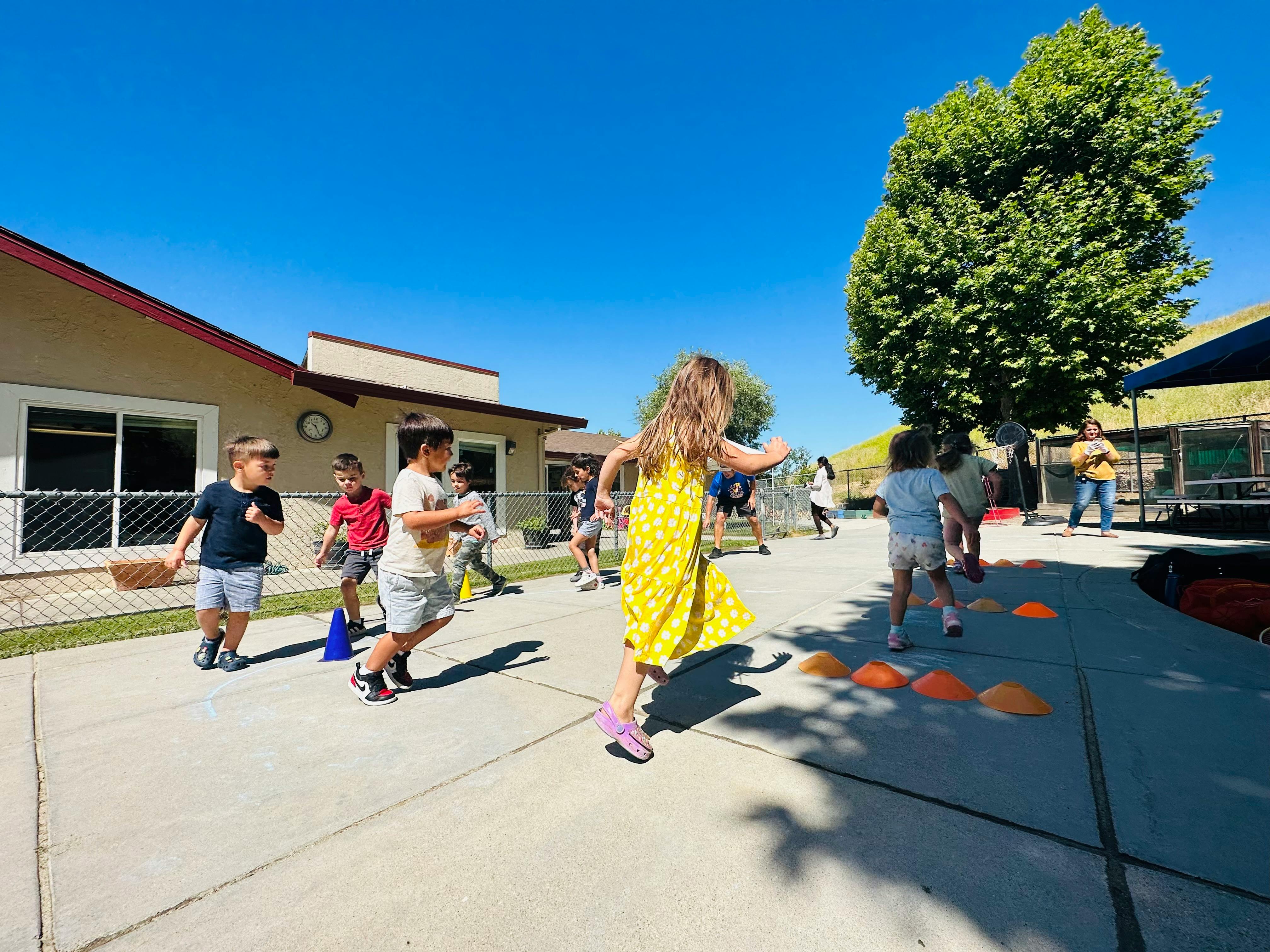 Children running outdoors on a paved area with buildings and a tree in the background