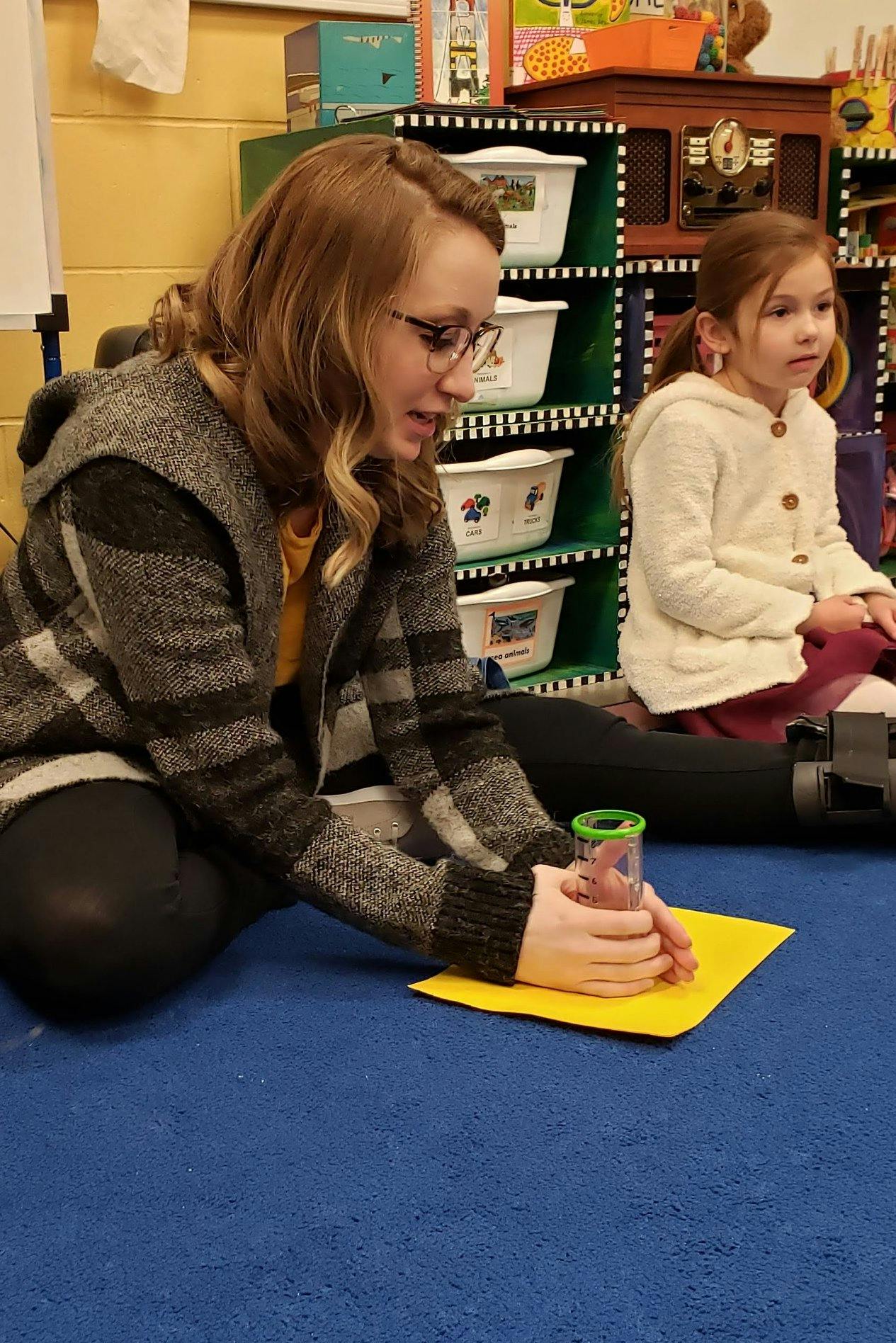Person holding a clear container over a yellow paper, child sitting nearby
