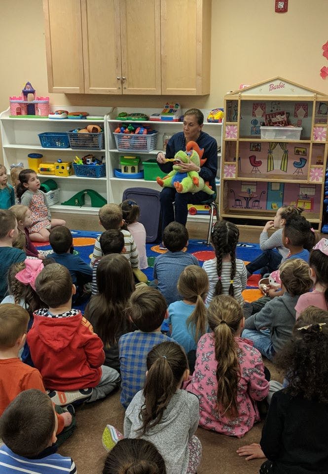 Person holding a stuffed animal and speaking to a group of children in a classroom