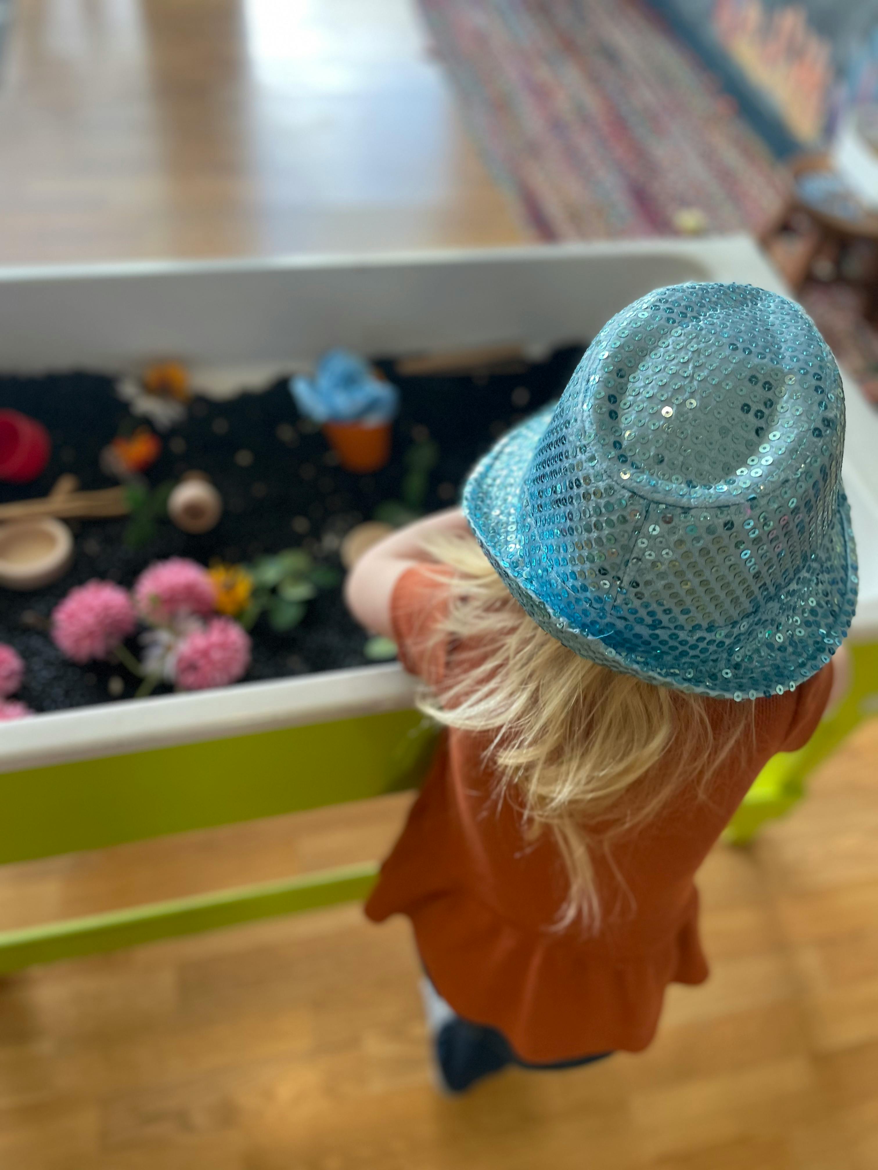 Child in a blue sequined hat looking into a sensory bin