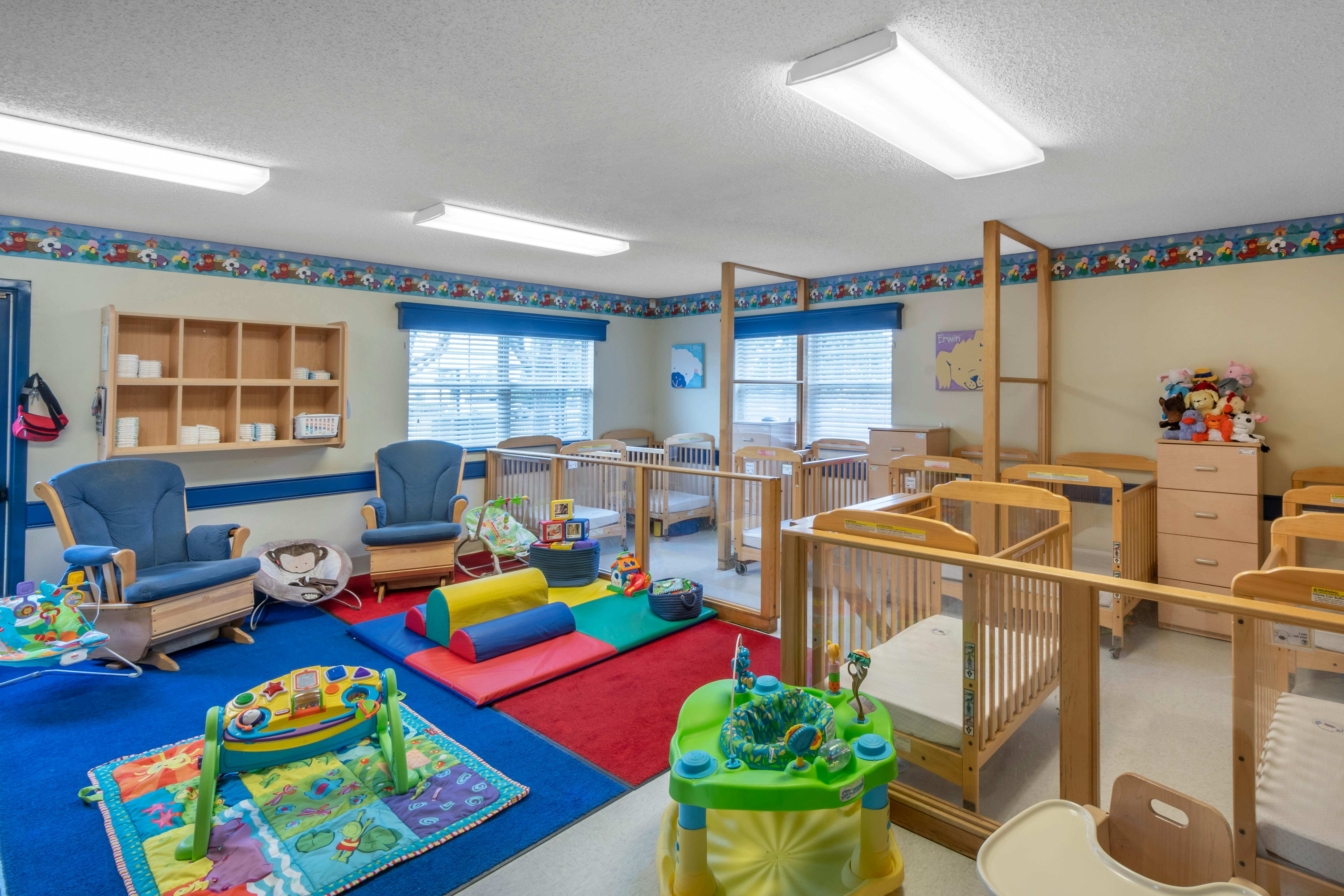 A colorful childcare room with cribs, play mats, and rocking chairs