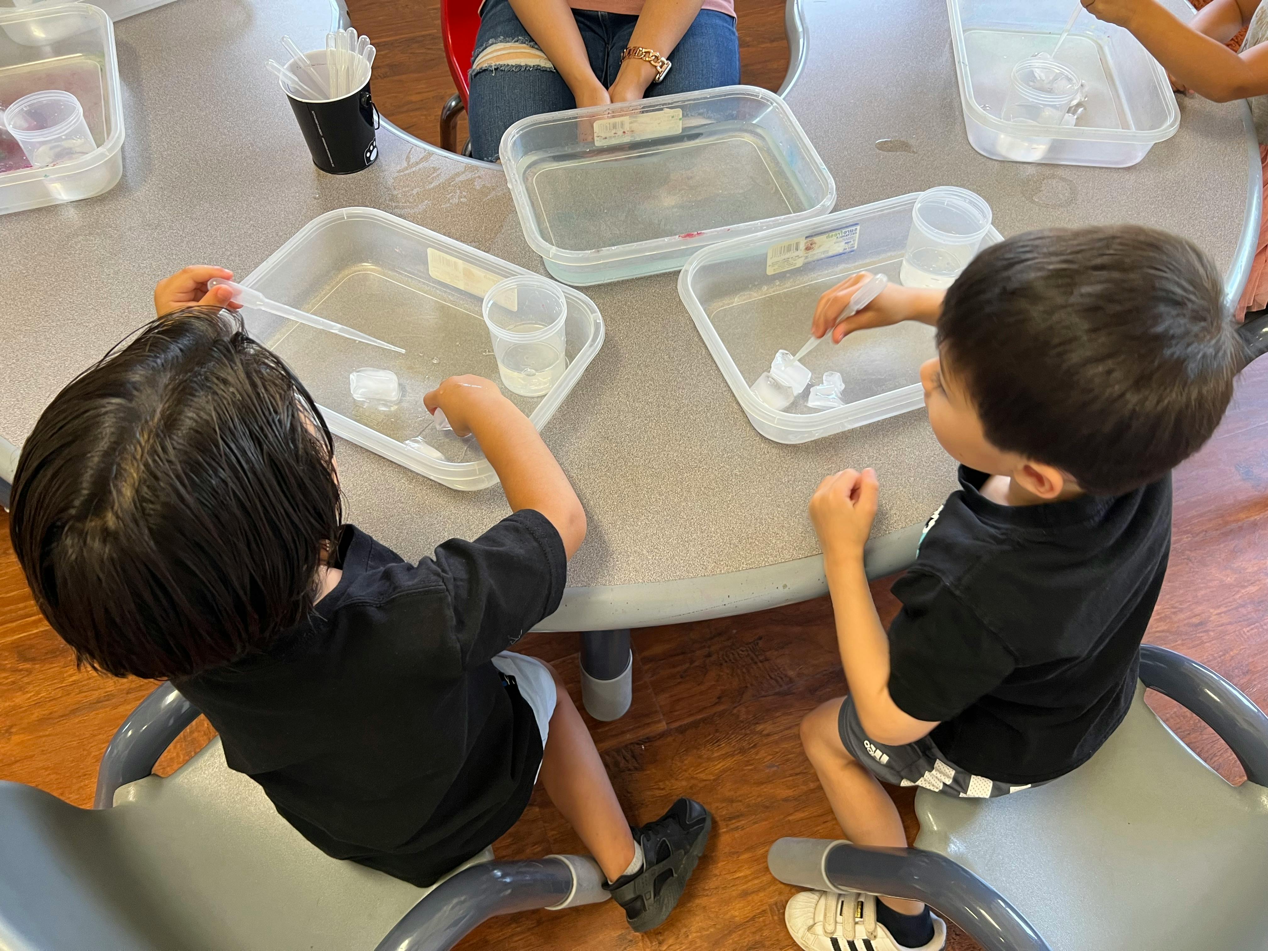 Two children at a table playing with water in plastic containers