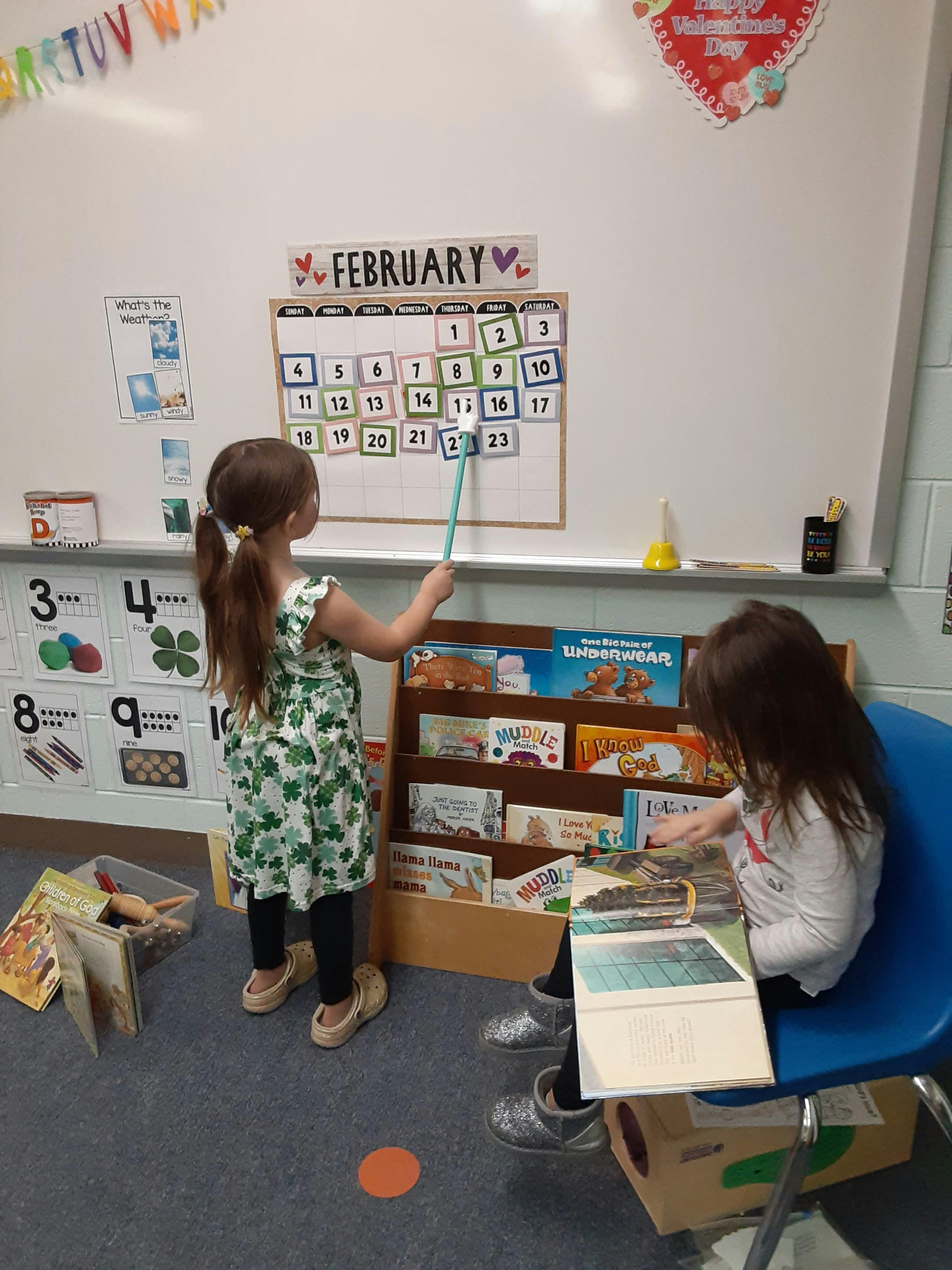Child pointing at a calendar on a whiteboard, another child reading a book