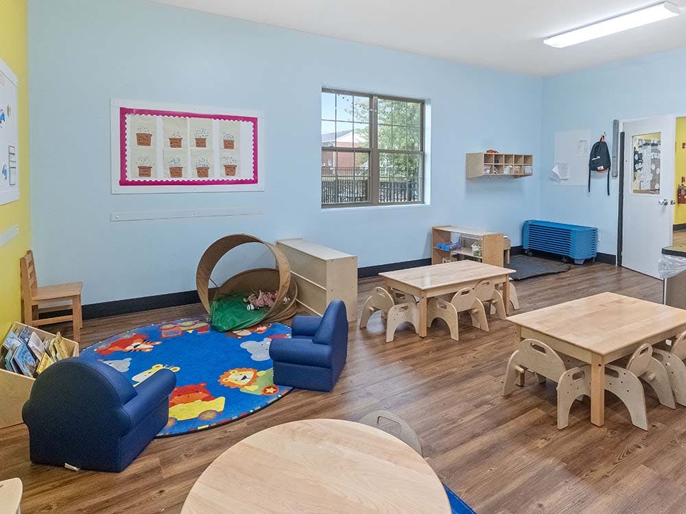 Classroom with small wooden tables and chairs, a blue rug, and play tunnel