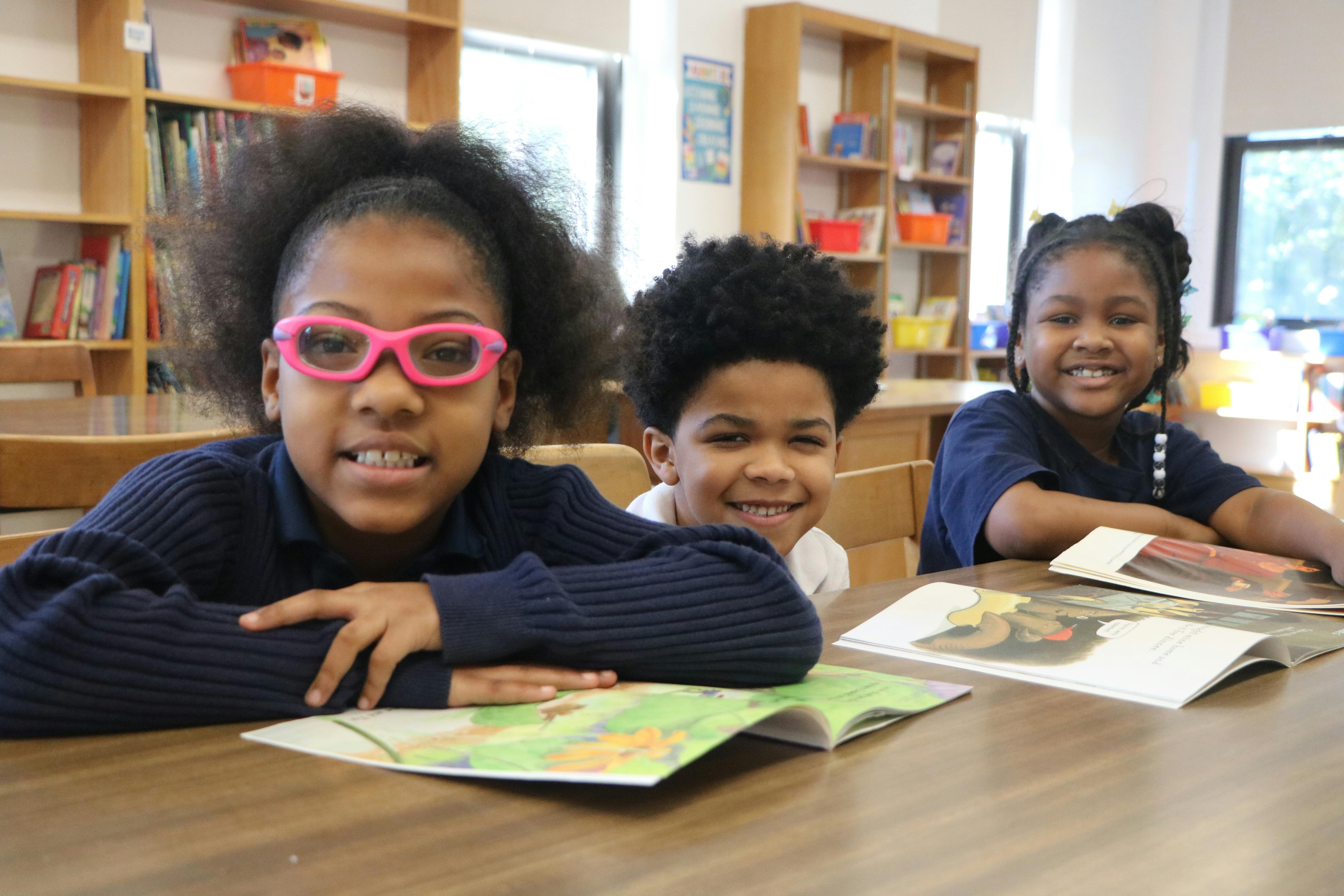 Three children sitting at a table with open books in a classroom