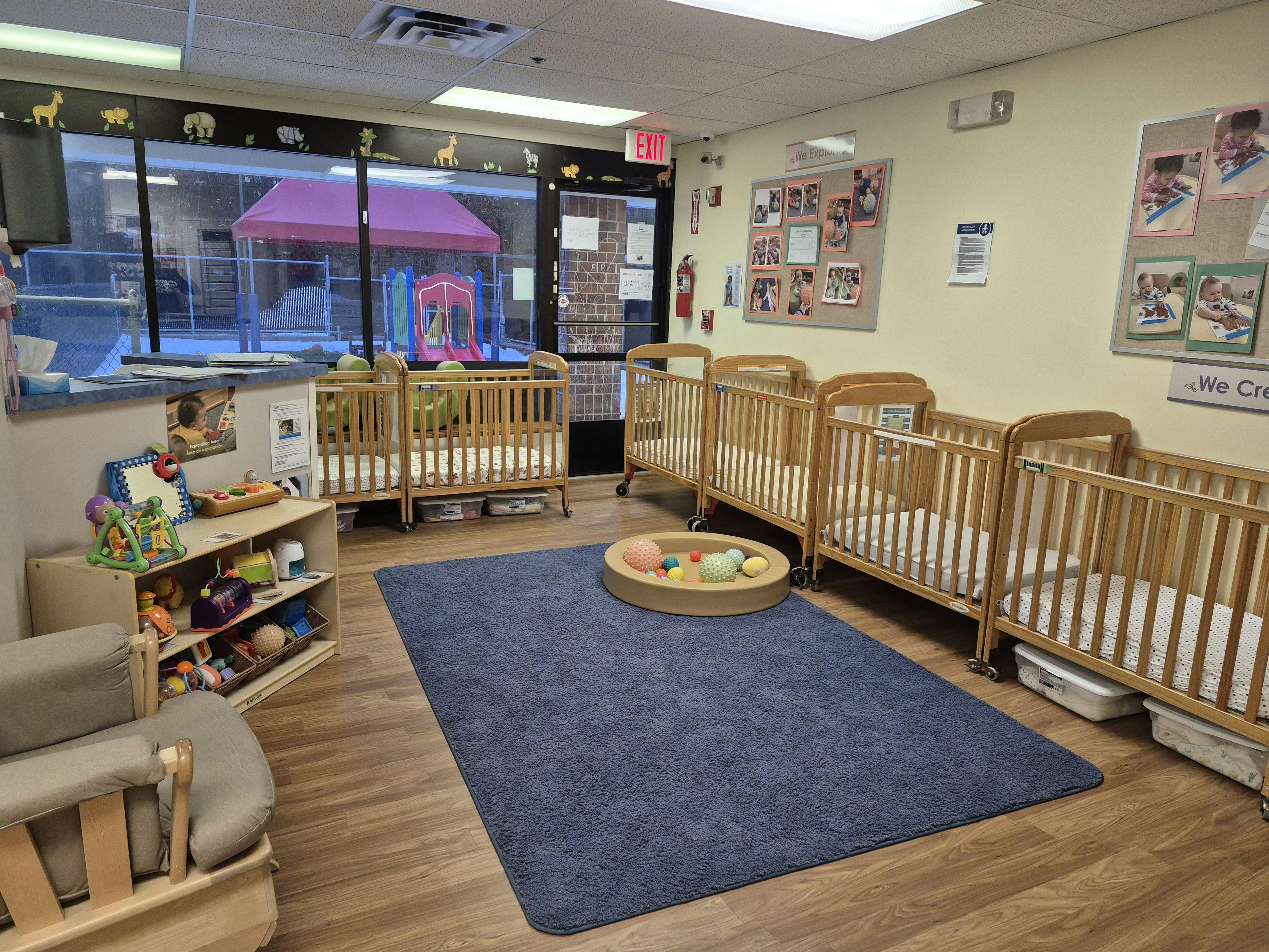 Cribs and play area in a daycare room, with an outdoor playground visible through the window