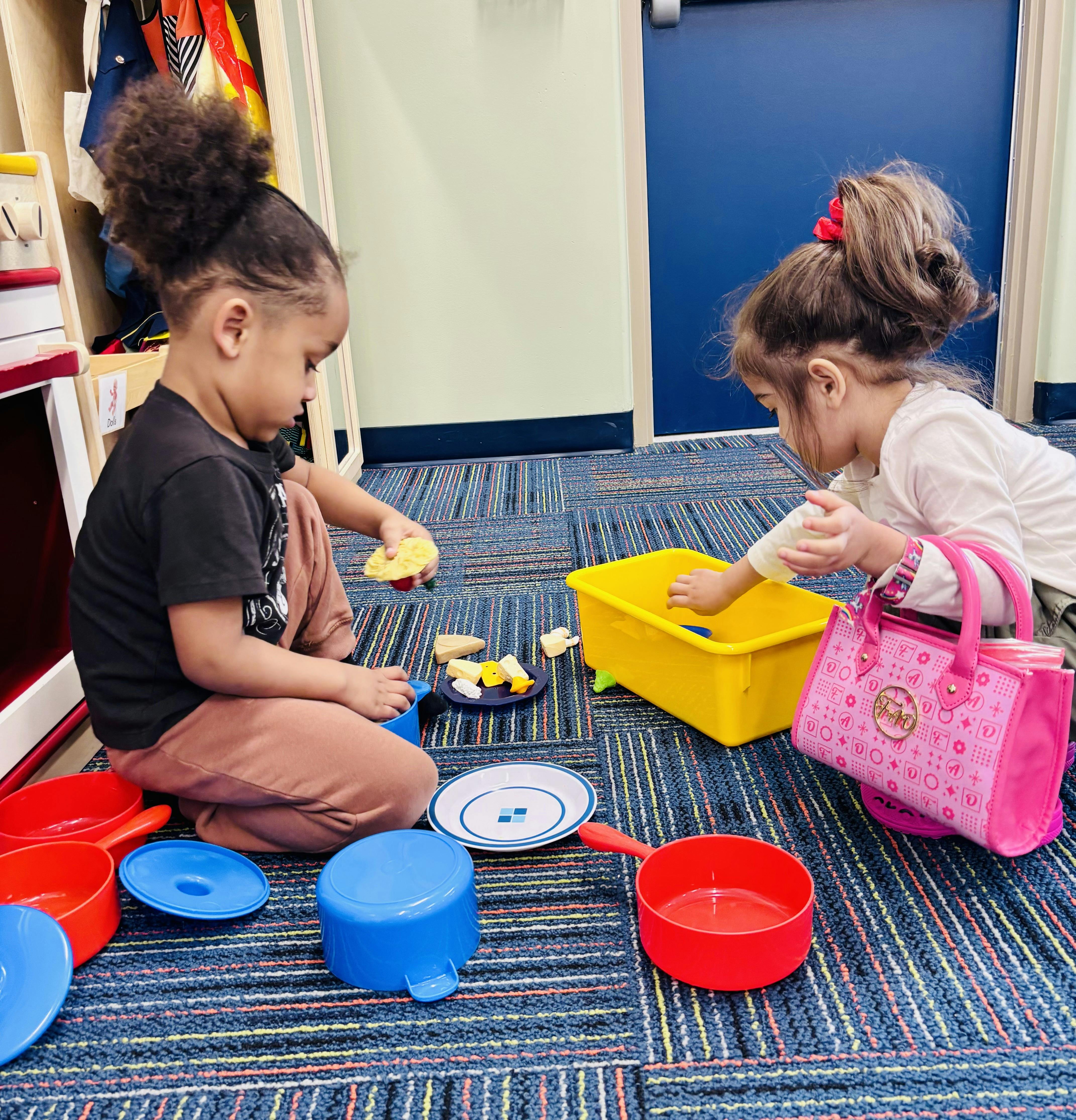 Two children playing with toy food and dishes on a classroom floor