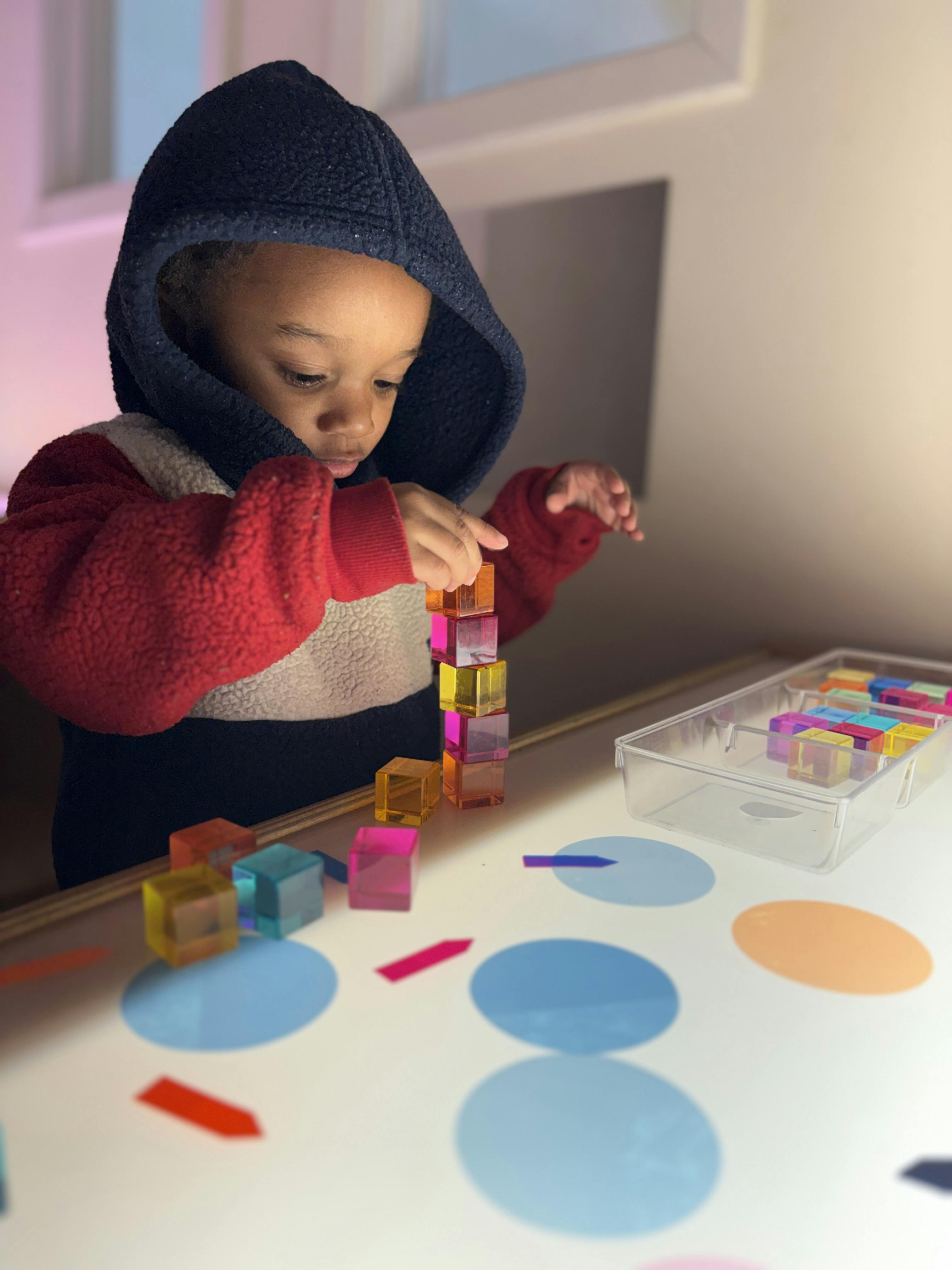 Child in a hooded sweatshirt stacking colorful translucent blocks