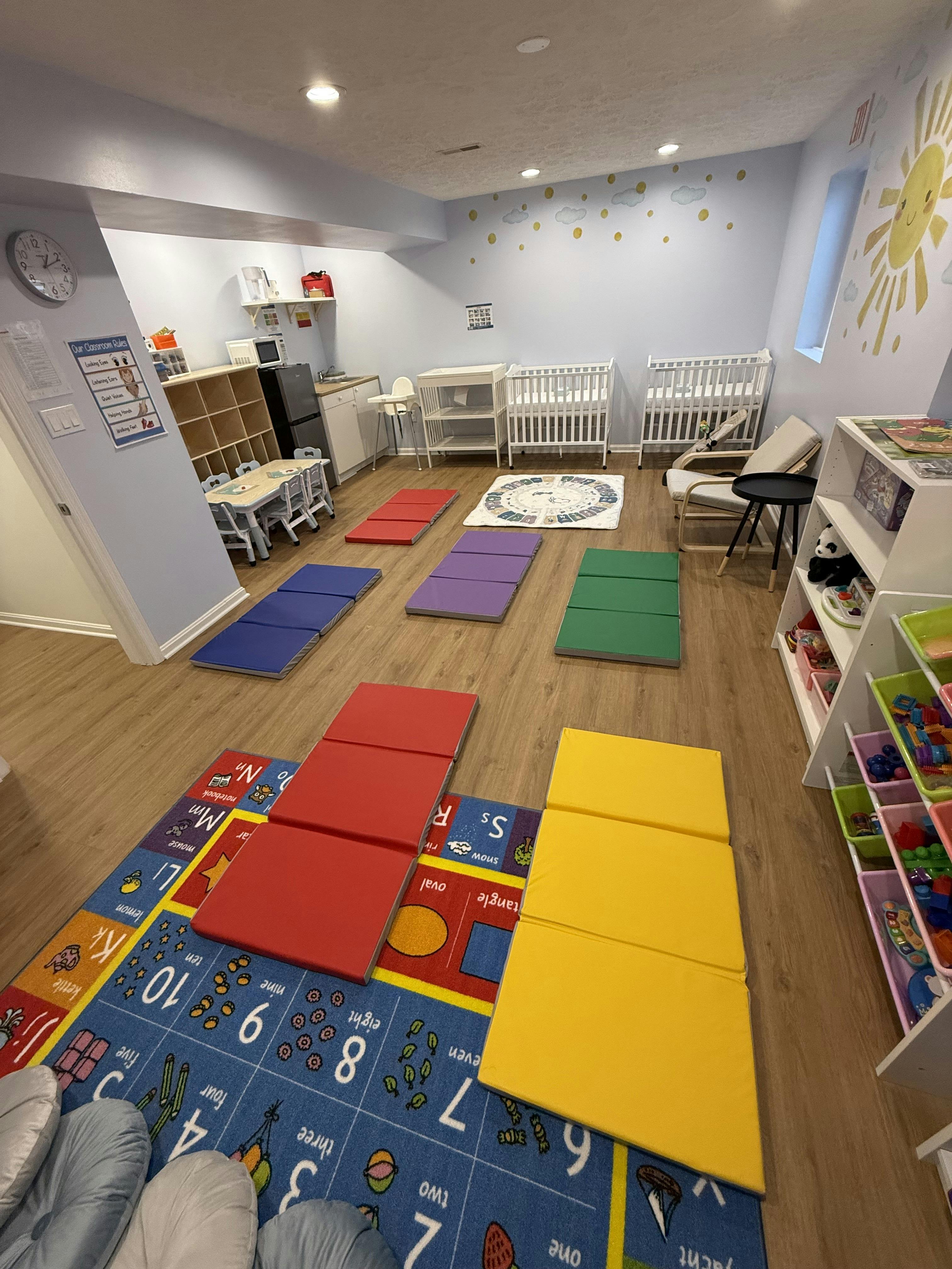 Daycare room with colorful mats on the floor and cribs in the background