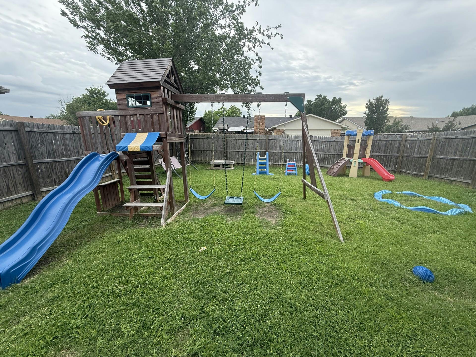 Playground in a backyard with a wooden playset, blue slide, and swings