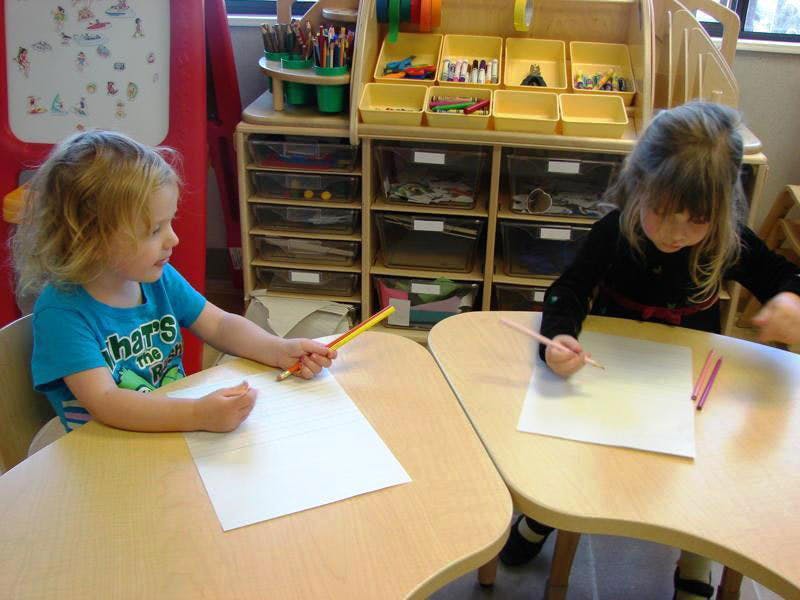Two children drawing at separate desks in a classroom