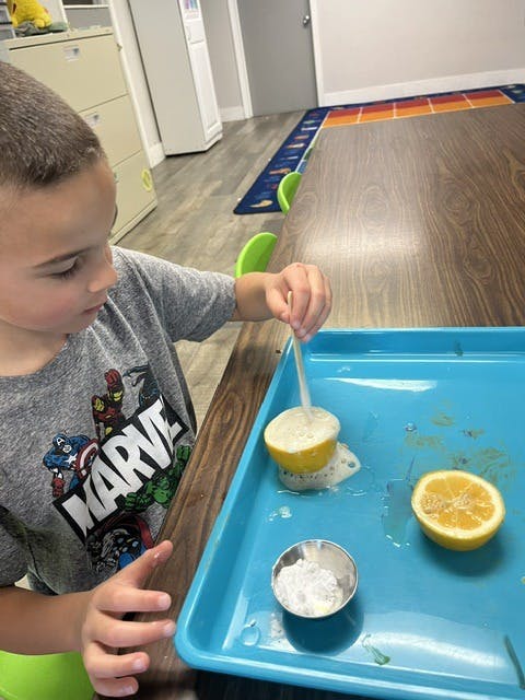 Child stirring a halved lemon with a stick, creating foam, on a blue tray