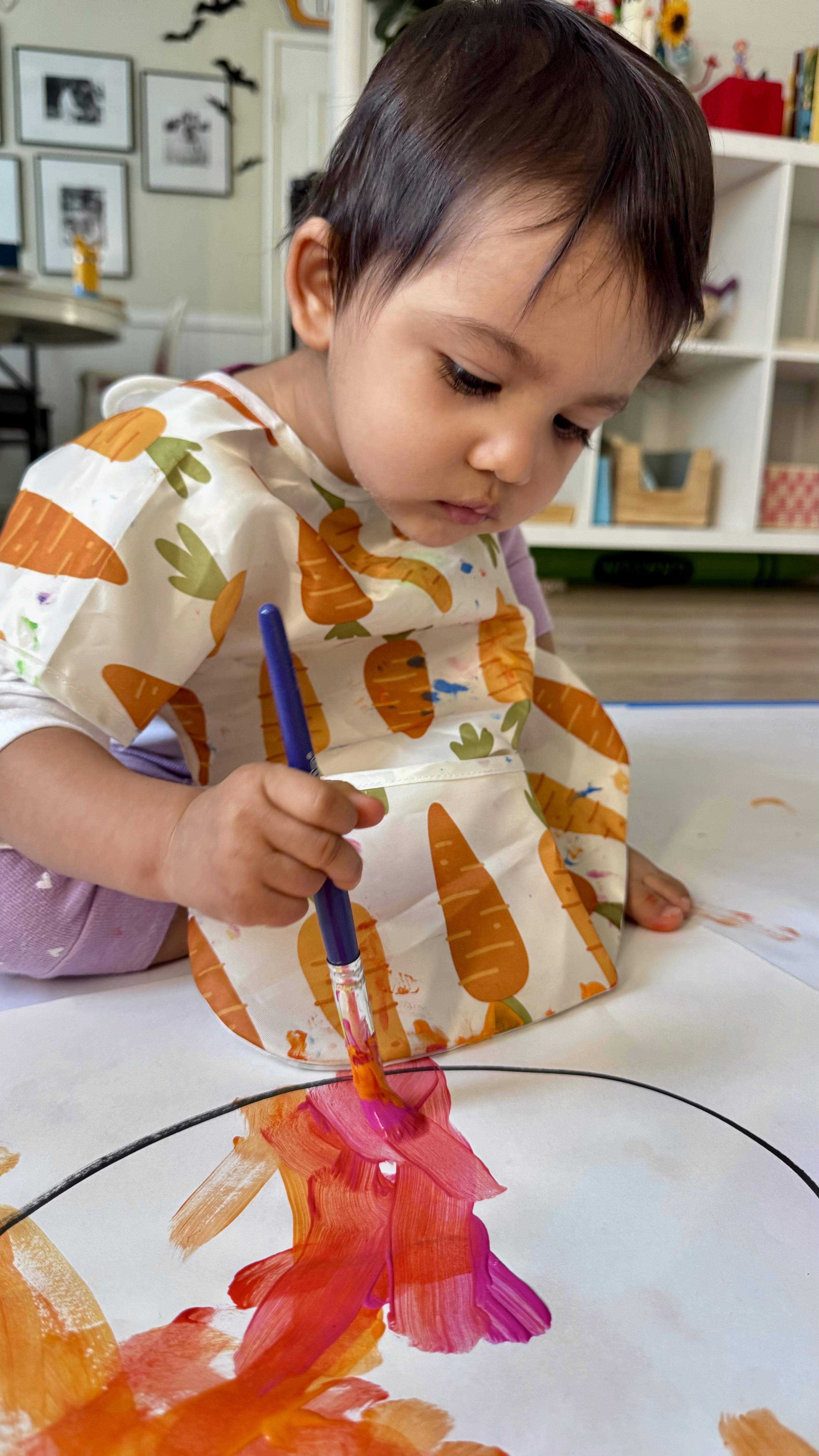 Child in a carrot-patterned bib painting with a brush