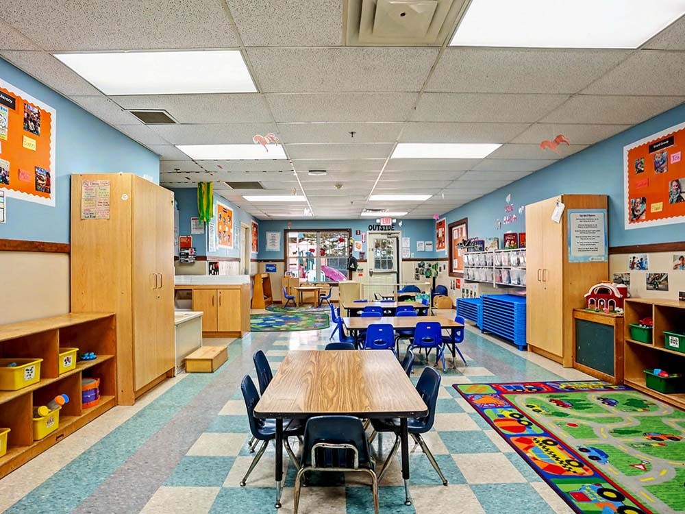 Classroom with small tables and chairs, shelves, and a playmat