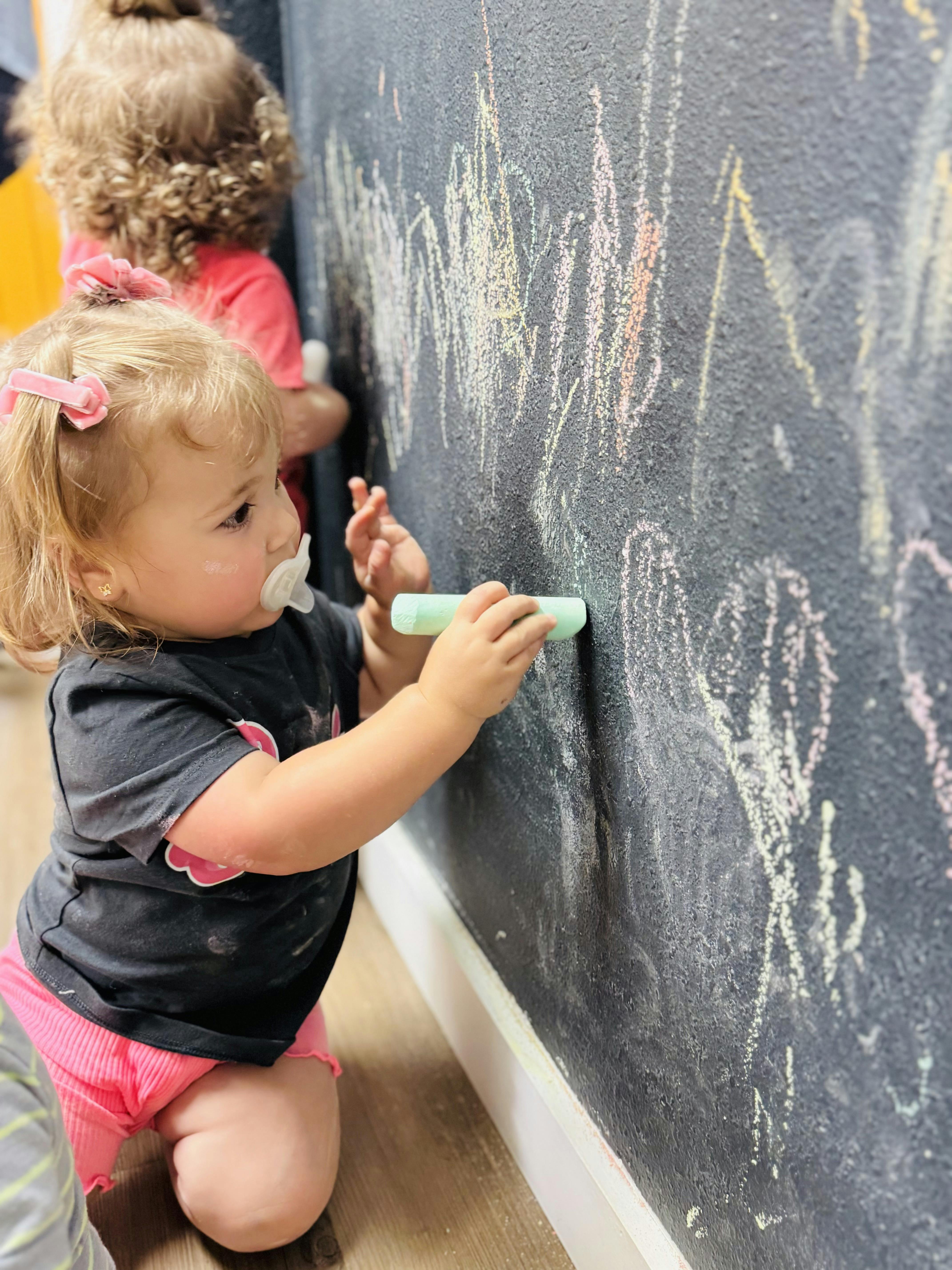 Child with pacifier drawing on a chalkboard with chalk