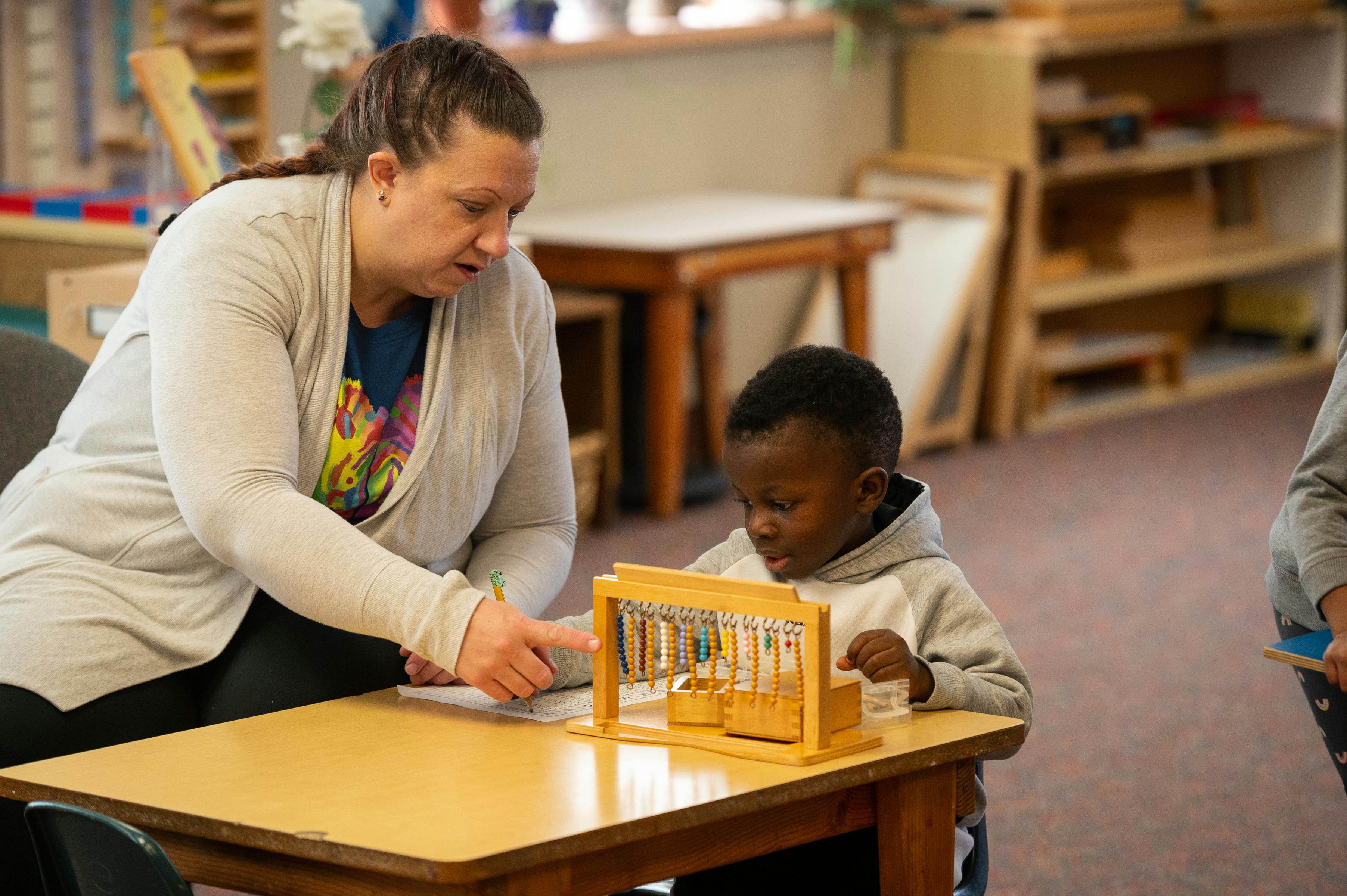 Person pointing at a child playing with a wooden toy in a classroom
