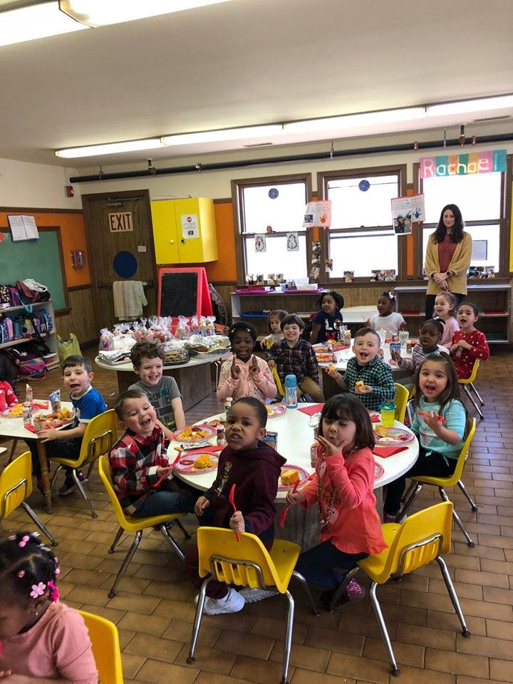 Children sitting at tables in a classroom with an adult standing in the background