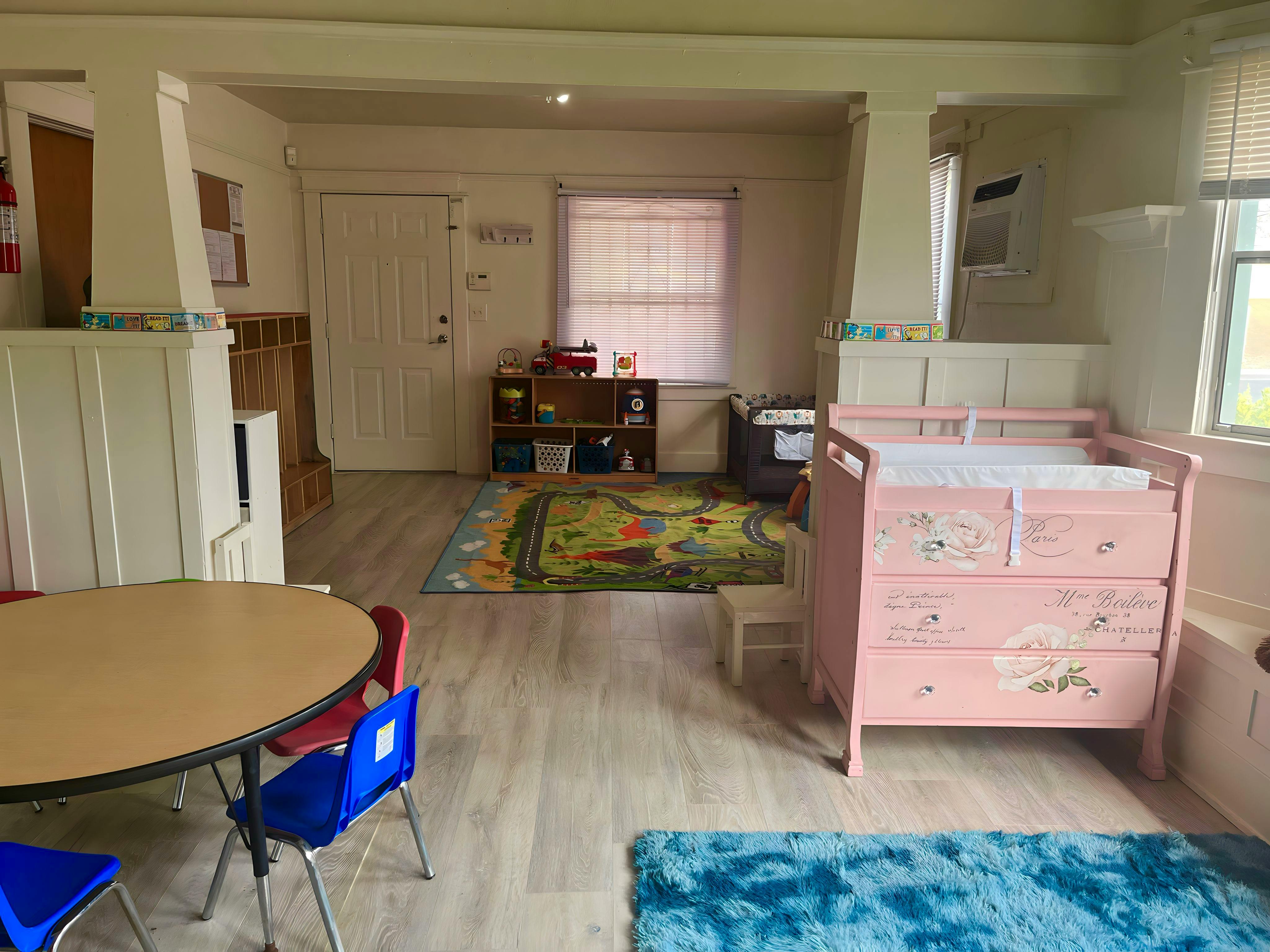 A room with a small table and chairs, a play rug, and a pink changing table