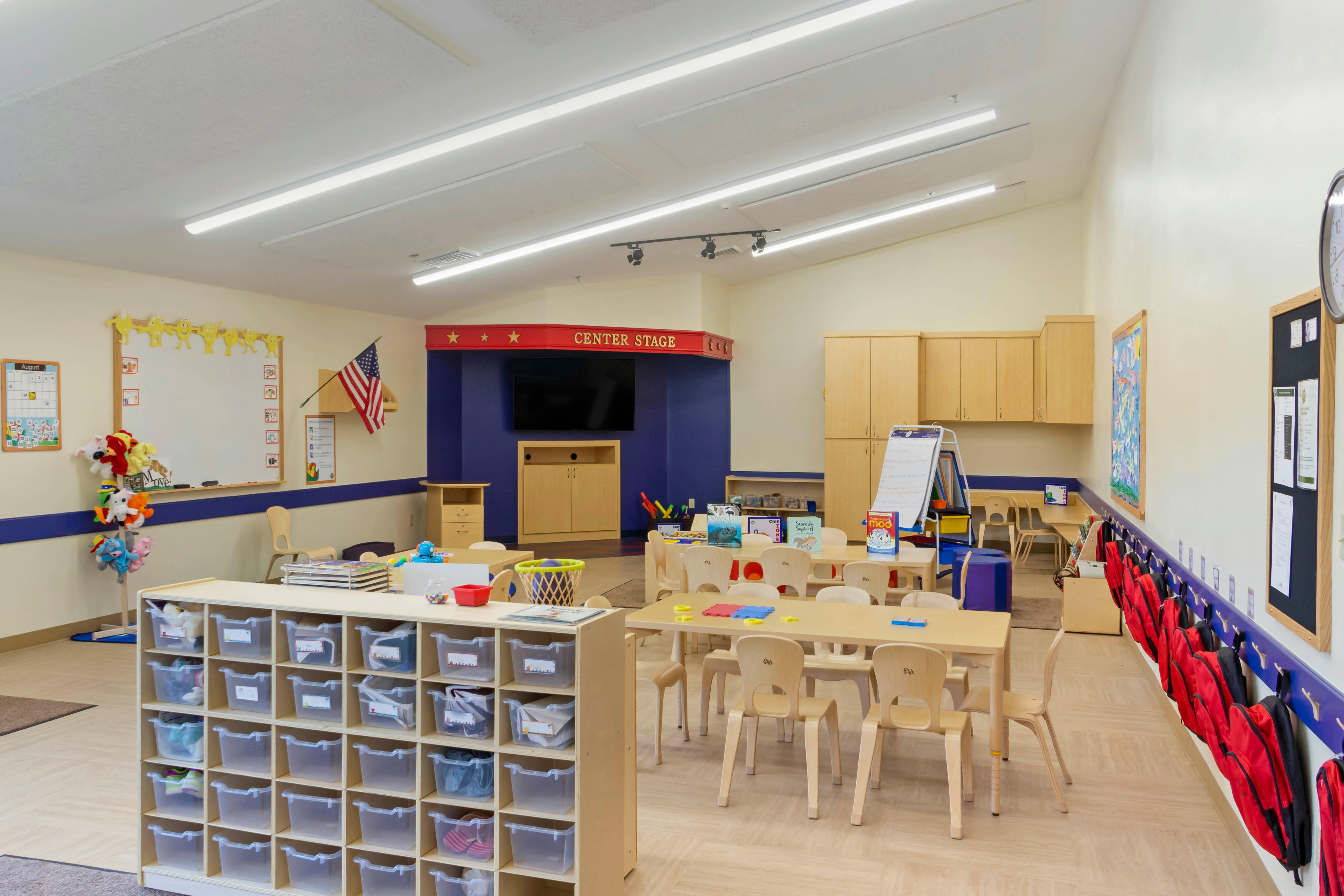 An empty classroom with small tables and chairs, cubbies, and a center stage