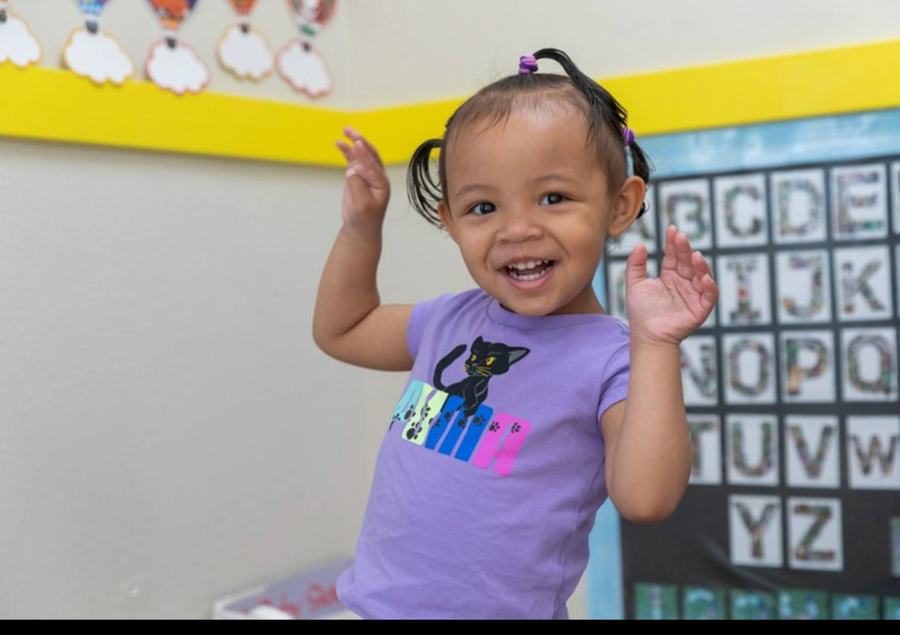 Child in a classroom with pigtails and purple shirt, hands raised