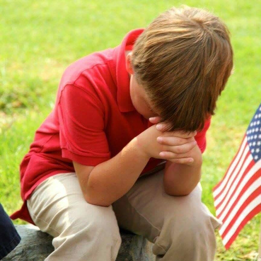 Child with head down and hands clasped, next to an American flag