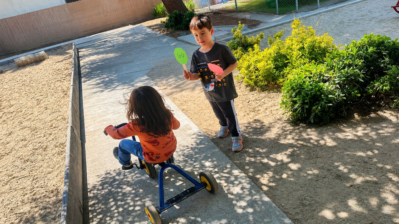 Child riding a tricycle while another holds a Go sign on a playground