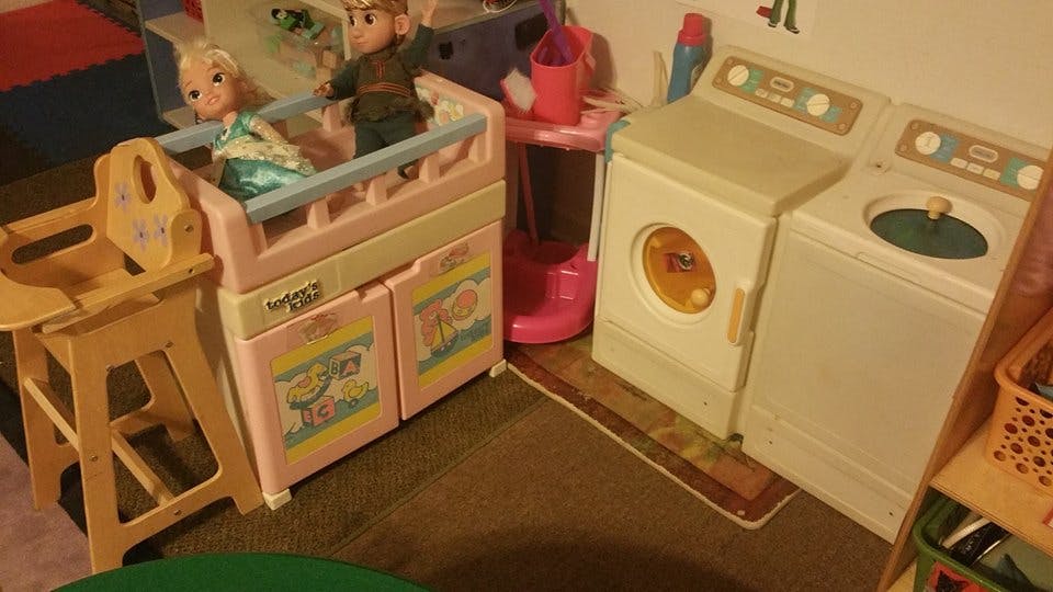 Dolls in a toy crib next to a toy high chair and toy washer and dryer