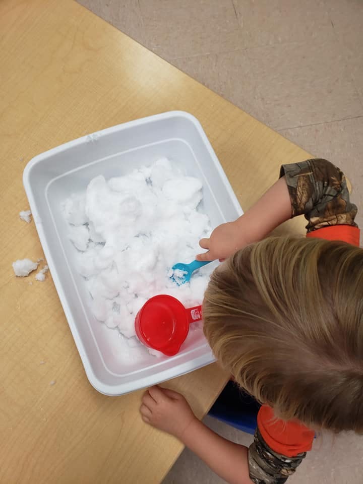 Child playing with snow in a bin with a red scoop and blue shovel