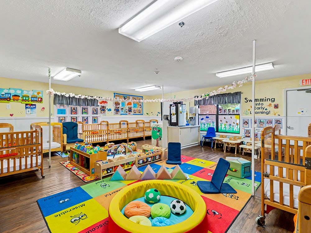 A colorful classroom with cribs, play mats, and small tables
