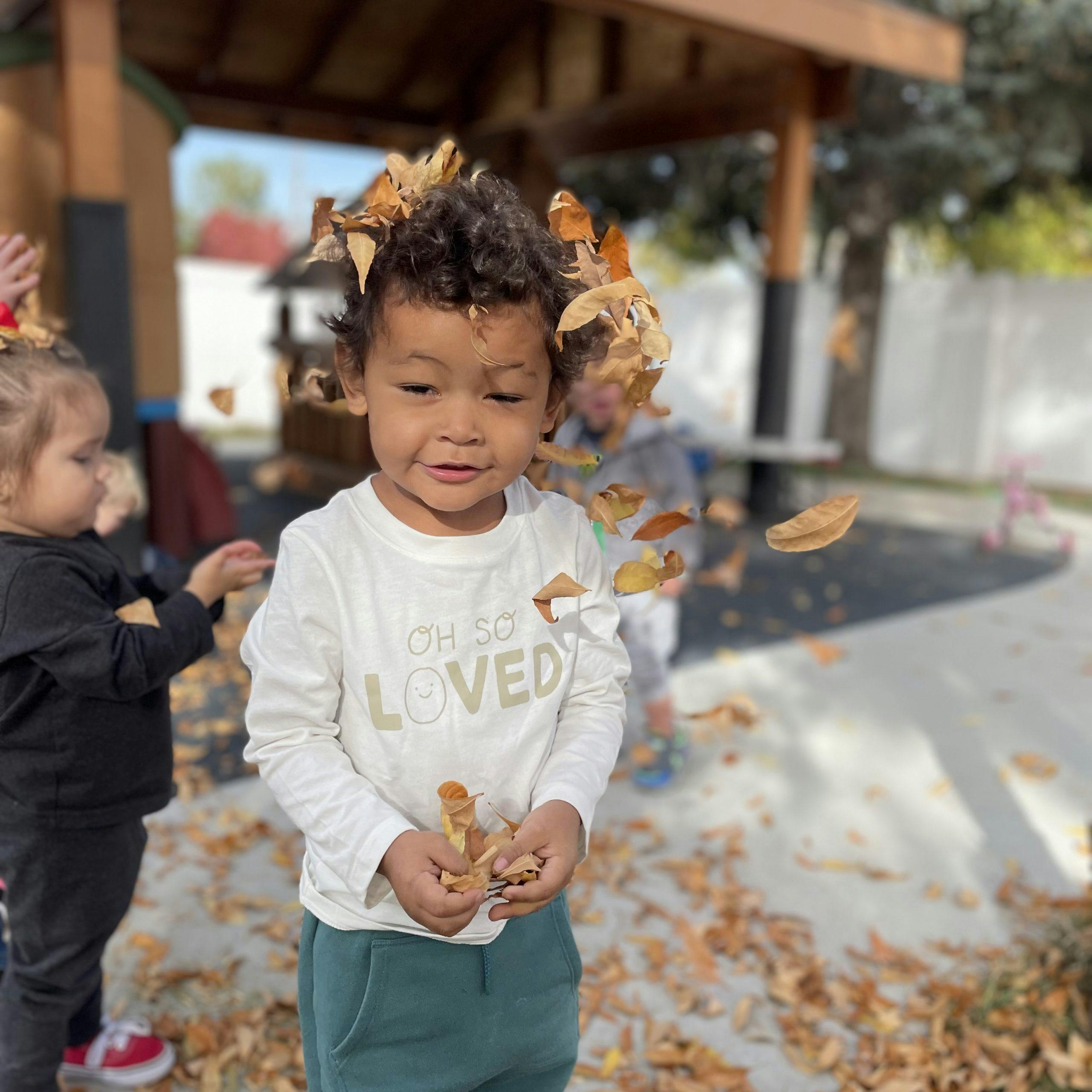 Child standing outside with fallen leaves on their head and in their hands