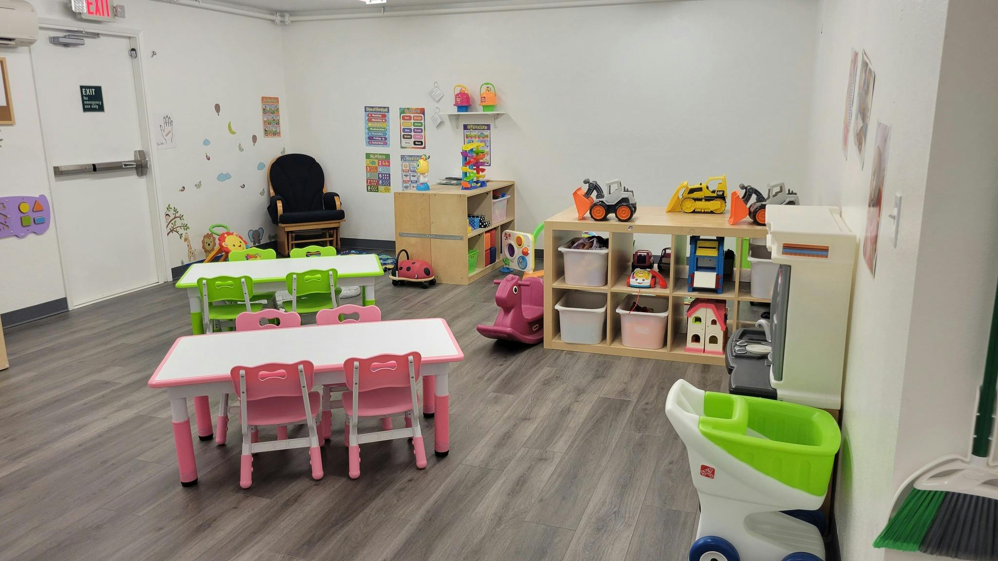 Classroom with small pink and green tables and chairs, shelves of toys