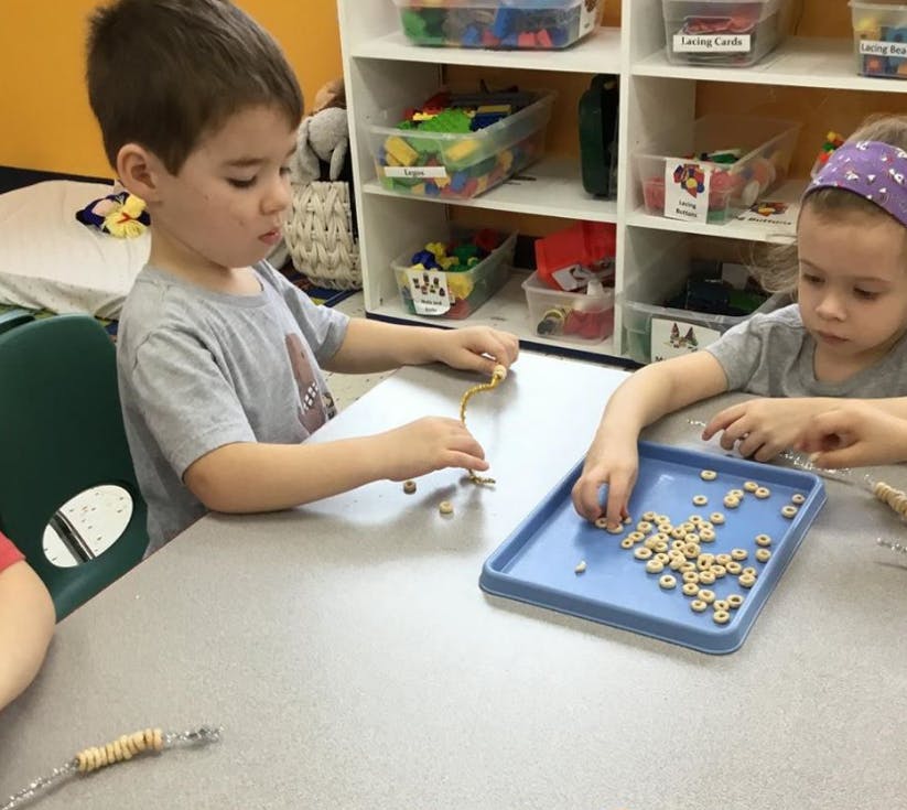 Two children at a table stringing cereal pieces onto pipe cleaners