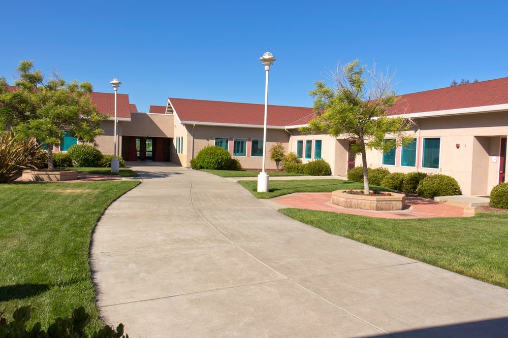 Building with red roof and light-colored walls, surrounded by green grass and trees
