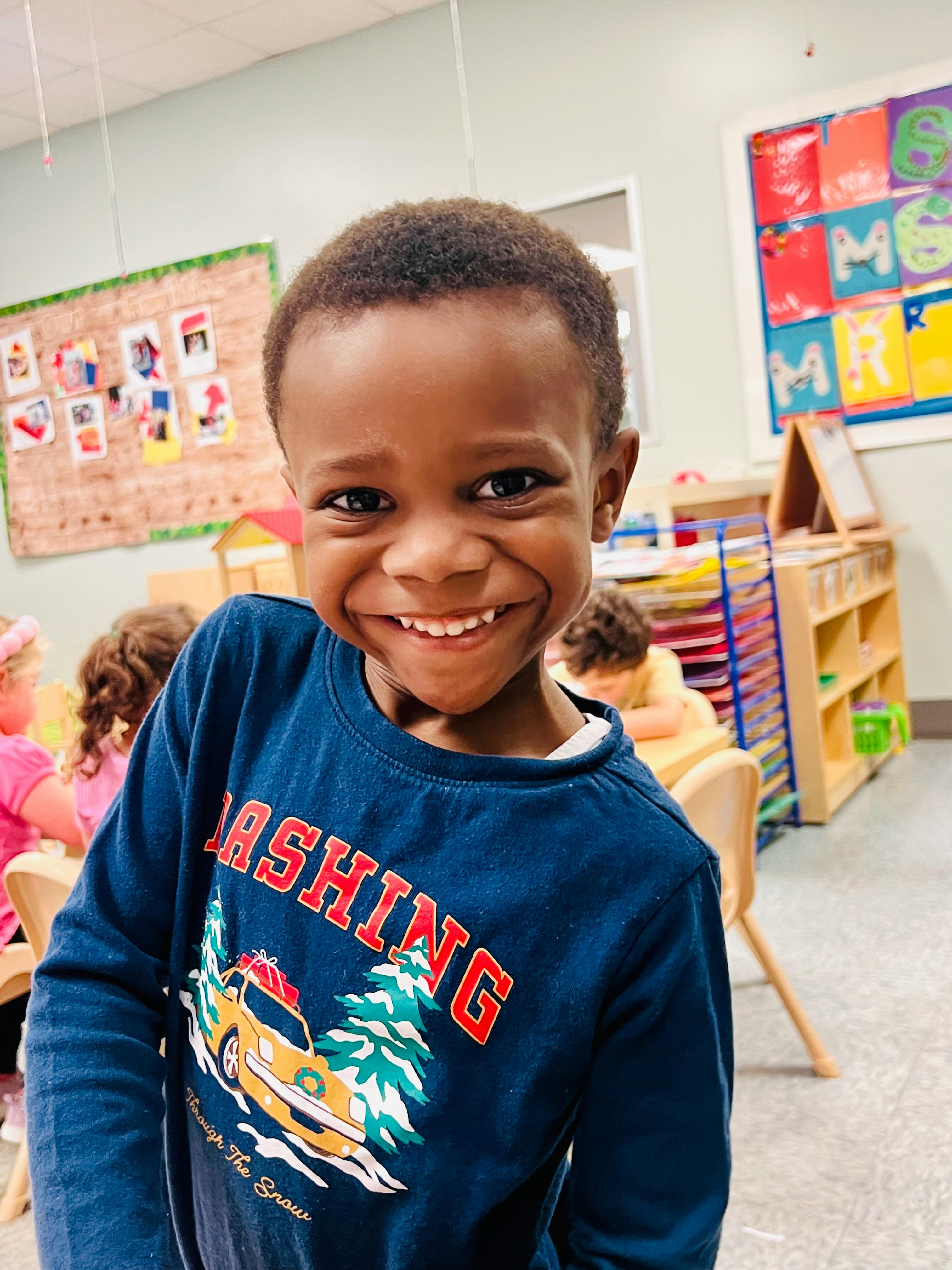 Child smiling widely in a classroom, wearing a navy blue shirt with a car graphic